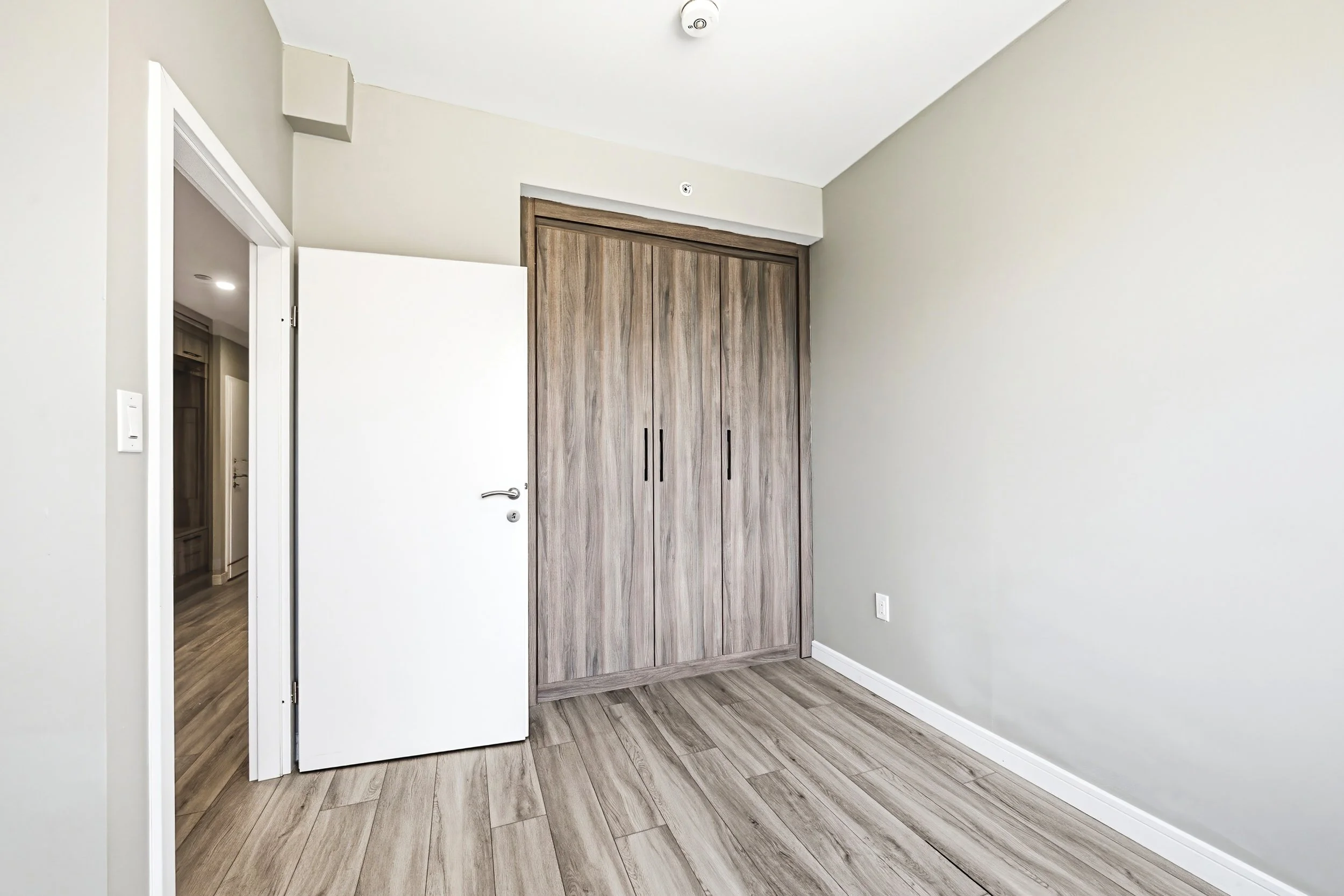 A room with light gray walls, wood-look flooring, an open white door, and a large wooden closet with sliding doors.