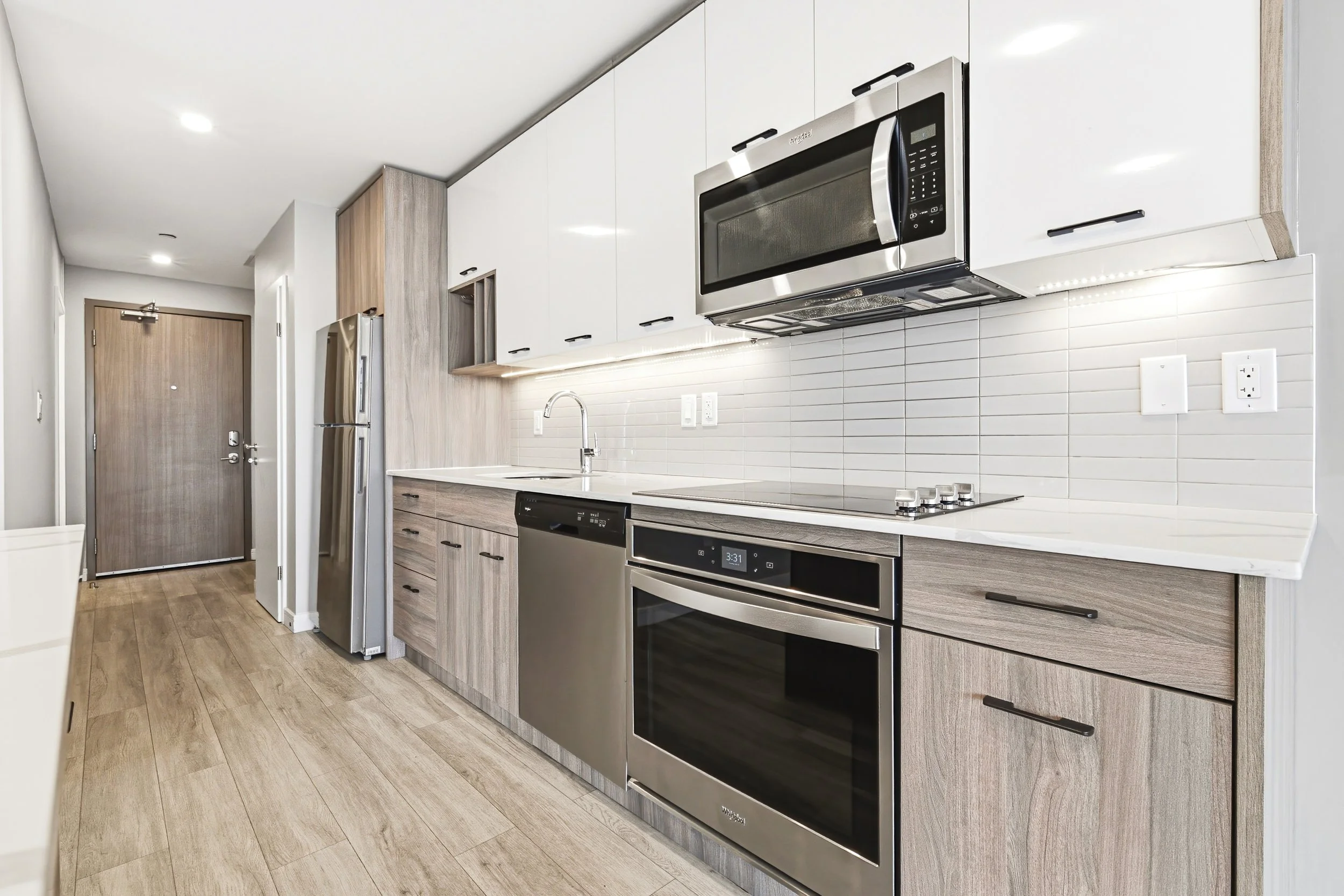 Modern kitchen with wooden cabinets, stainless steel microwave and refrigerator, oven, stovetop, white tile backsplash, and wood flooring.