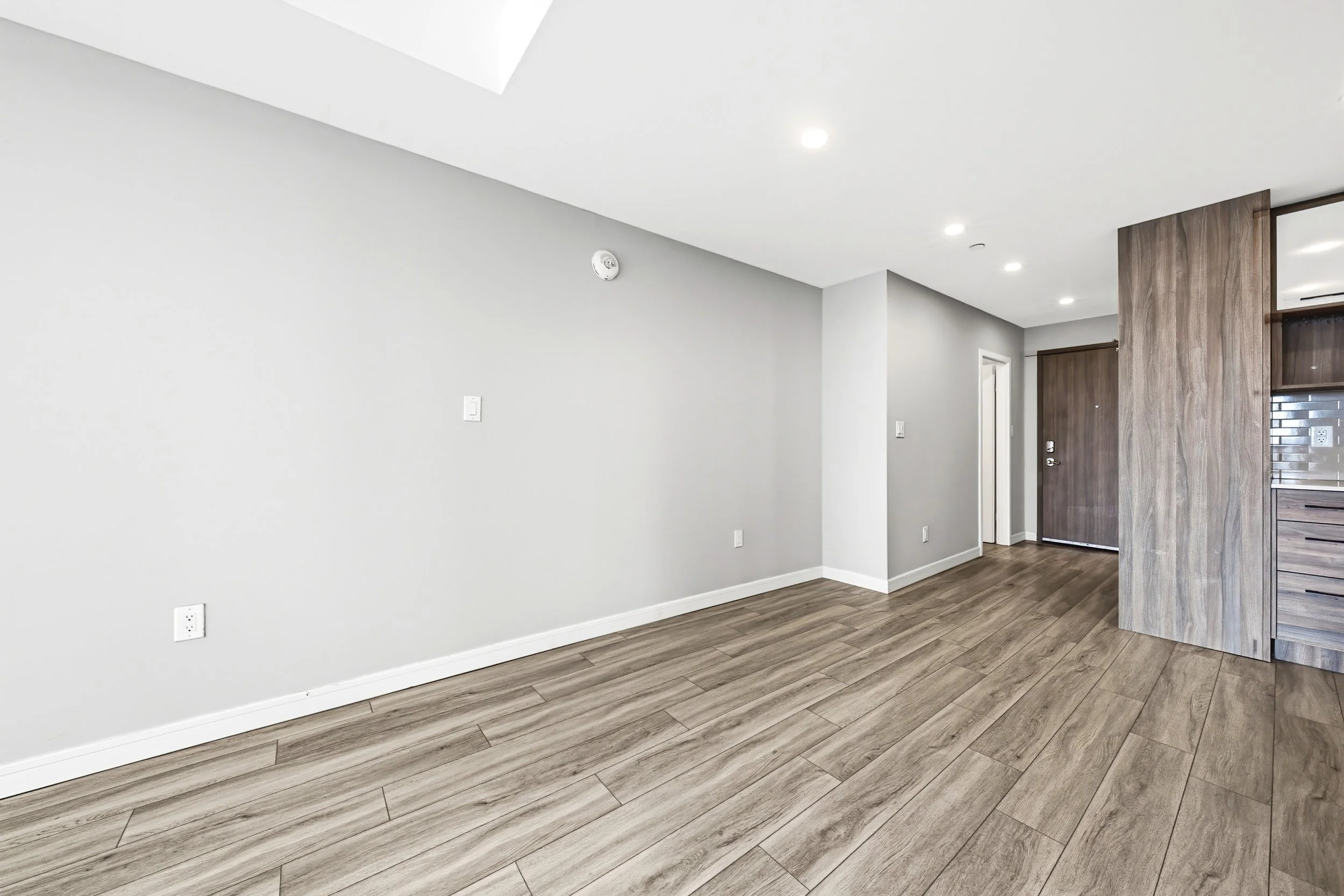 Empty living room with white walls, hardwood flooring, and a partial view of a wooden wall and door.