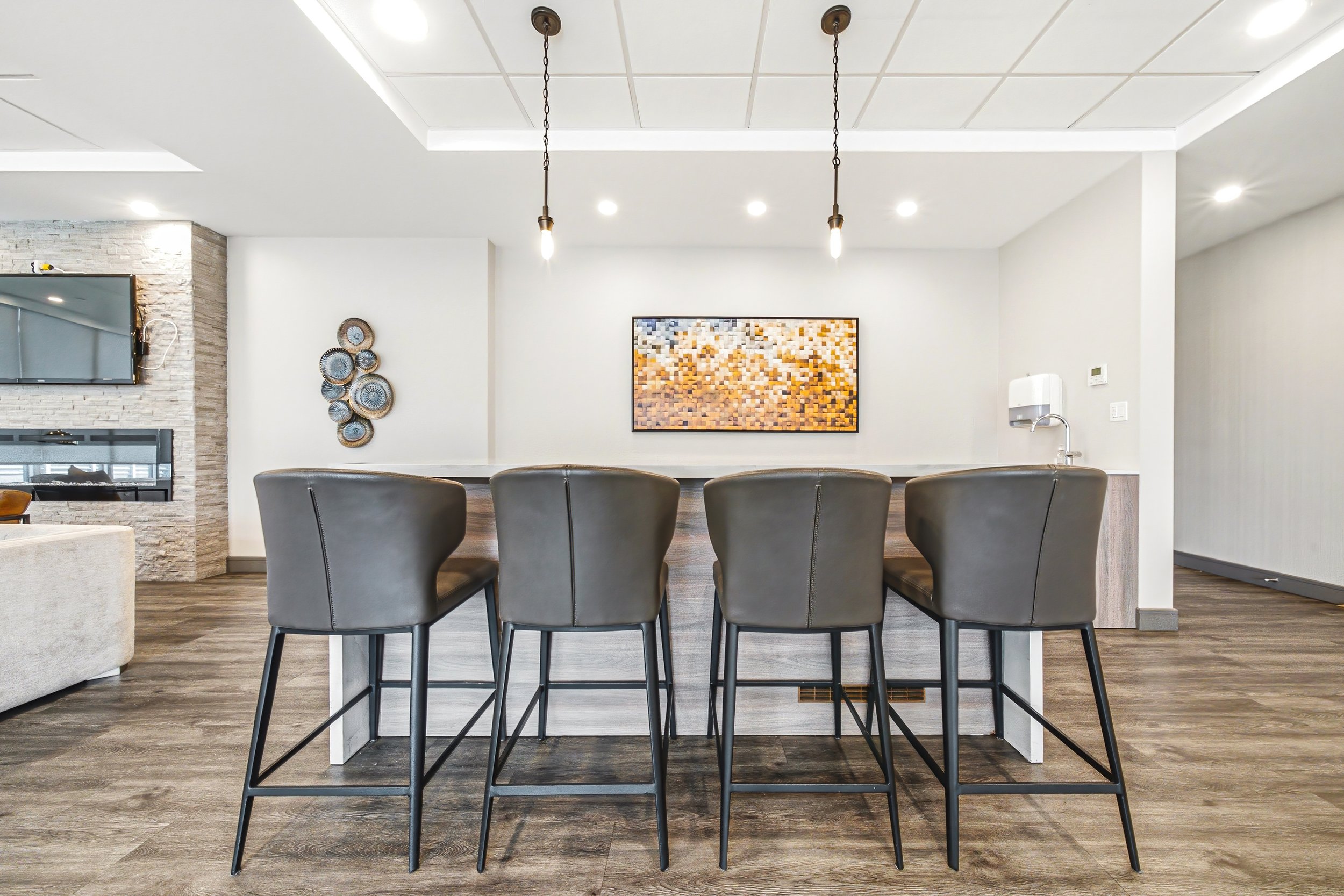 Modern dining or kitchen area with a white divider, five black chairs around a light-colored countertop, art on the wall, and a television on the left wall.