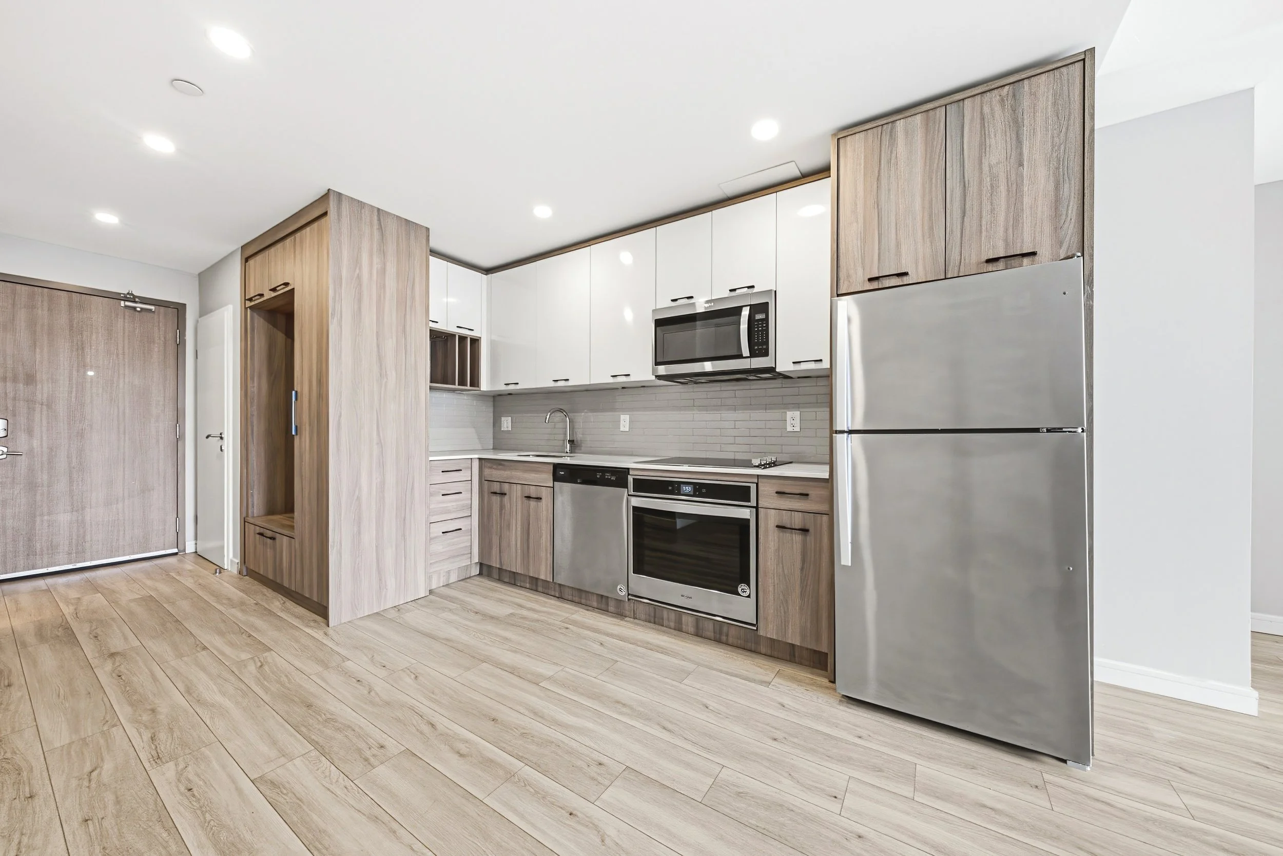Modern kitchen with wood and white cabinetry, stainless steel appliances including a refrigerator, microwave, dishwasher, and oven, and wood-look flooring.