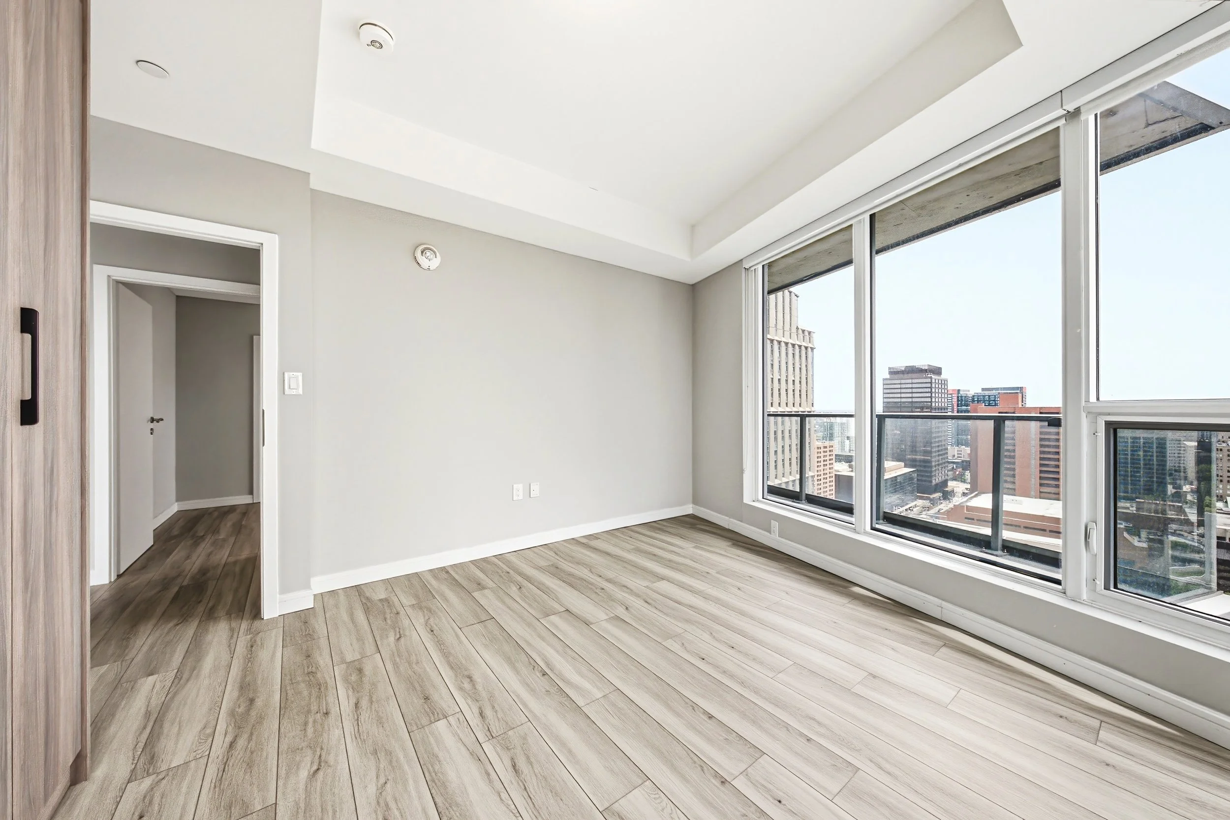 Empty room with large window showing city skyline, light wood flooring, white walls, and a ceiling with two smoke detectors.