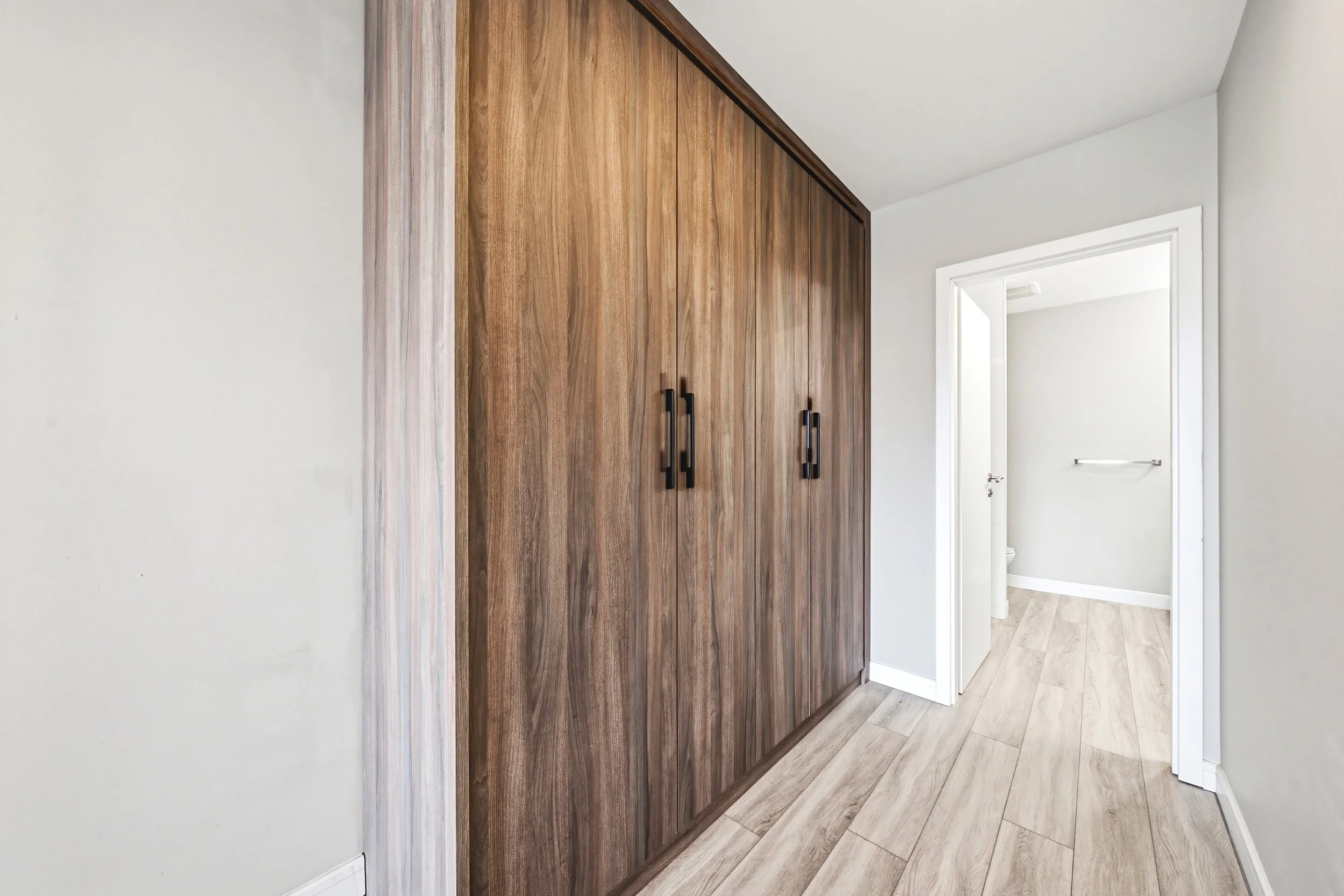 Inside a modern home, a large wooden closet with black handles is seen on a white wall next to a hallway with a door and a towel bar.