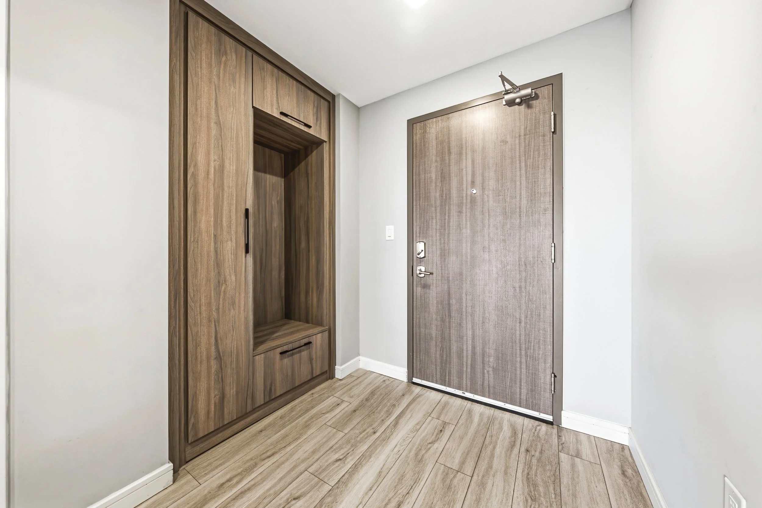 Interior view of an apartment entryway featuring a wooden closet with sliding doors and a small open storage space, next to a gray main door with a peephole and door hardware, light-colored walls, and wood-look flooring.