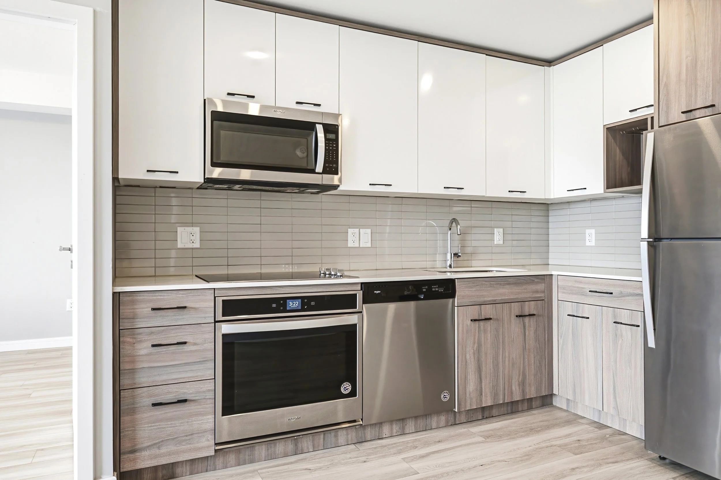 Modern kitchen with white upper cabinets, wood lower cabinets, a built-in microwave, stainless steel oven and dishwasher, a gray tiled backsplash, and a silver refrigerator.