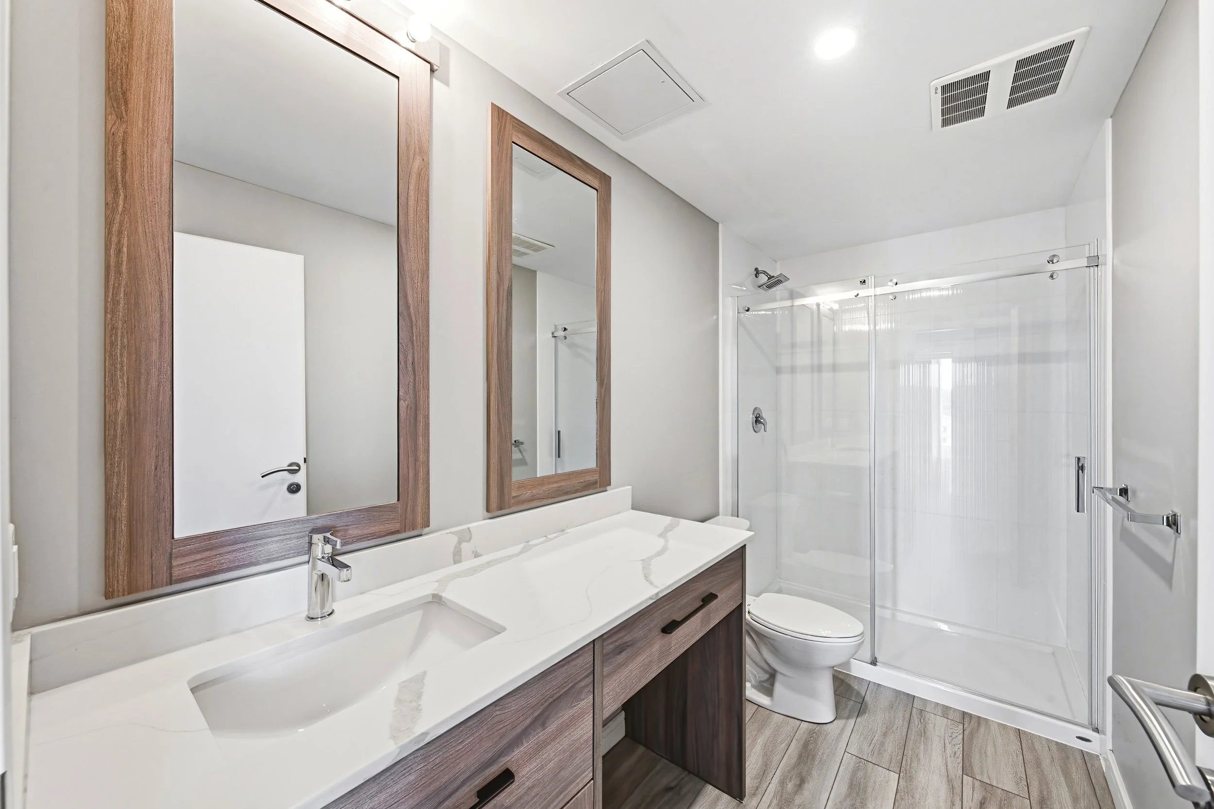 A modern bathroom with two mirrors framed in wood, a white marble countertop with two sinks, a wooden vanity, a toilet, and a glass shower enclosure with a showerhead, ceiling vents, and a door handle.