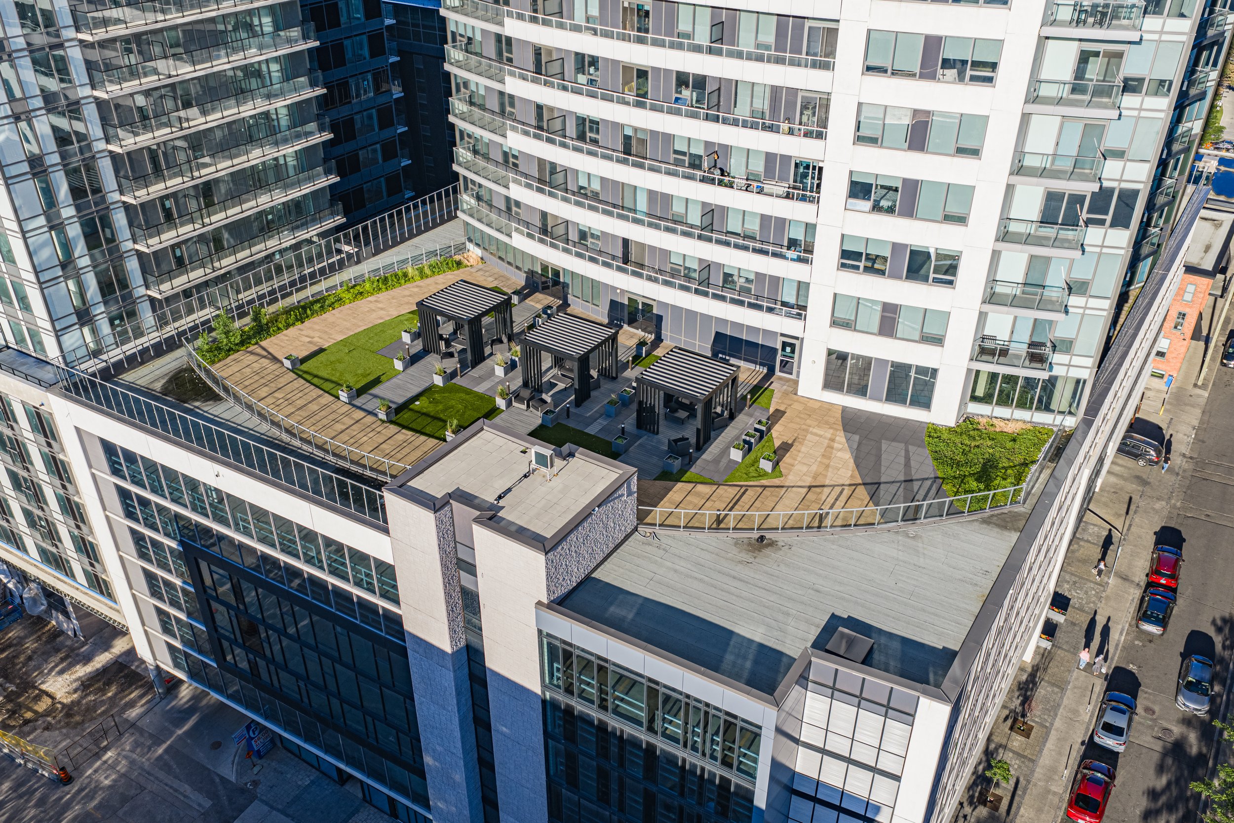 A high-rise apartment building with a rooftop terrace featuring outdoor seating areas, potted plants, and a small garden, with surrounding city streets and parked cars visible.