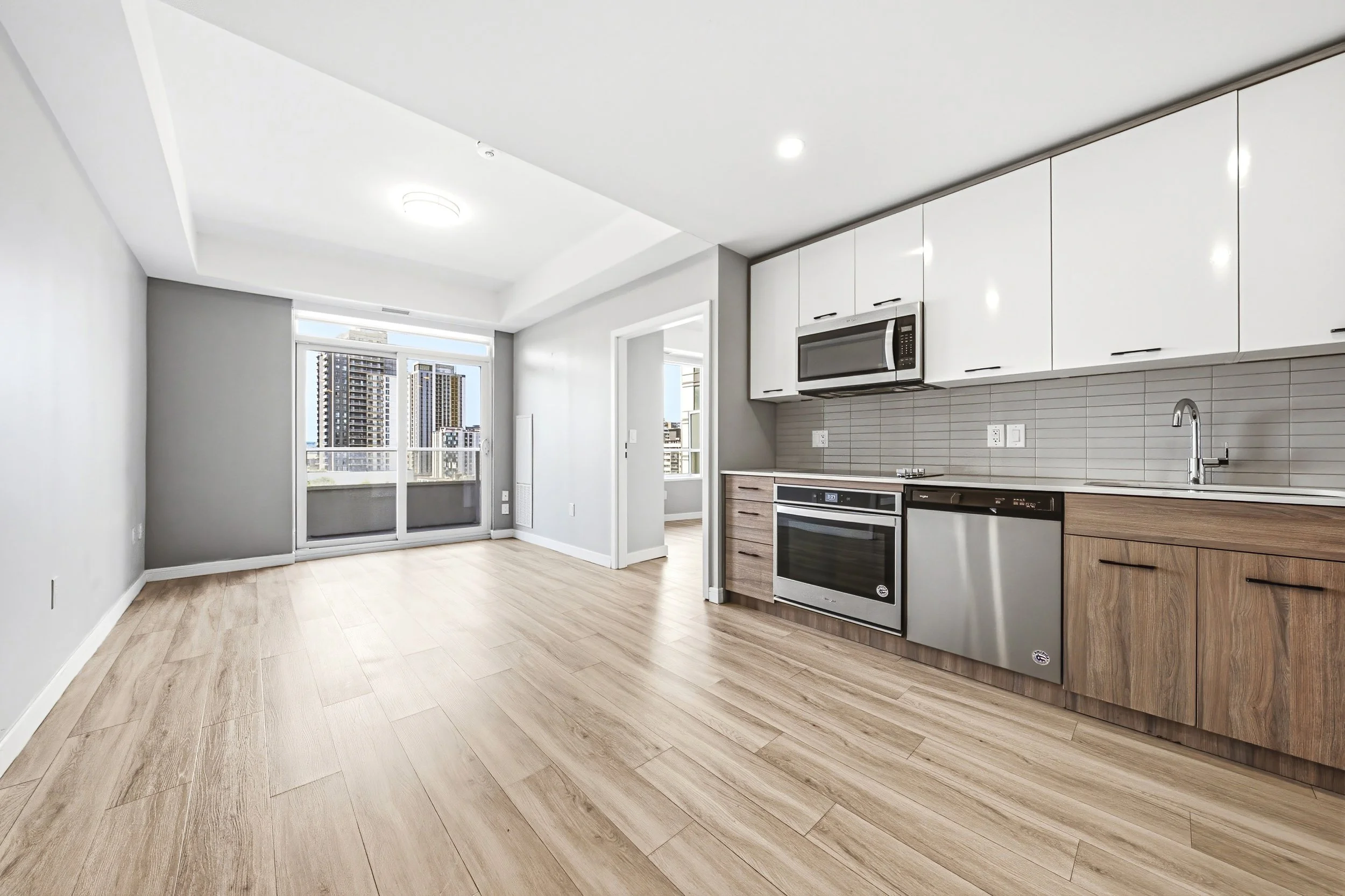 Modern kitchen with white cabinets, stainless steel microwave, oven and dishwasher, light wood flooring, and a balcony door with city view.