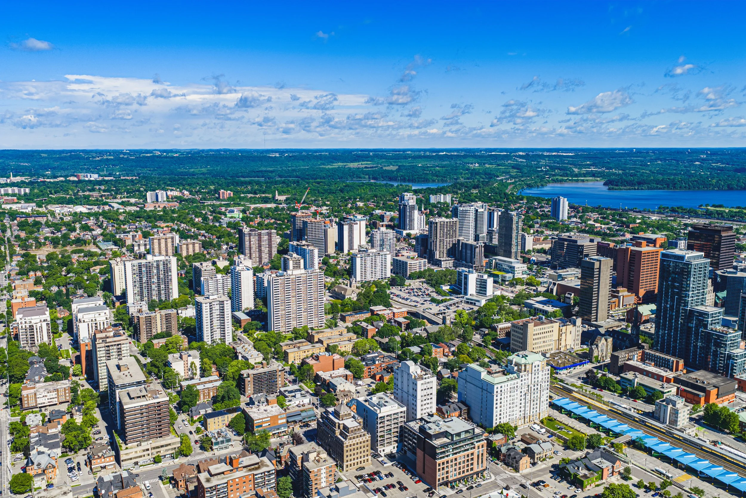 Aerial view of a city with high-rise buildings, green spaces, and a large body of water in the background under a partly cloudy sky.