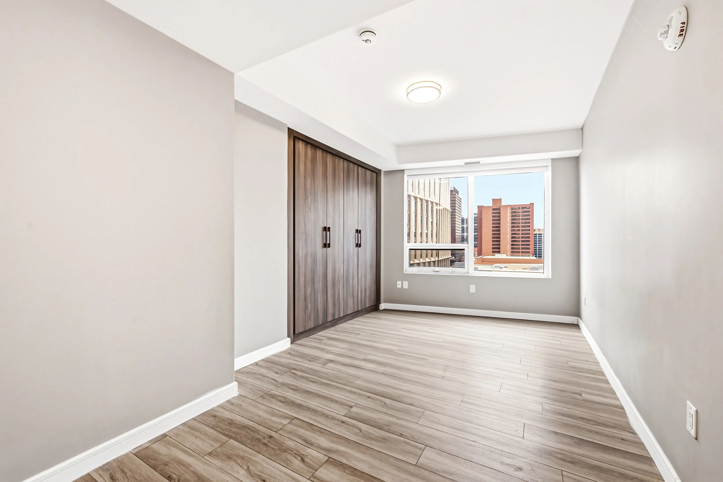 Empty room with light wood flooring, white walls, a built-in wooden closet, large window showing city buildings, and ceiling lights.