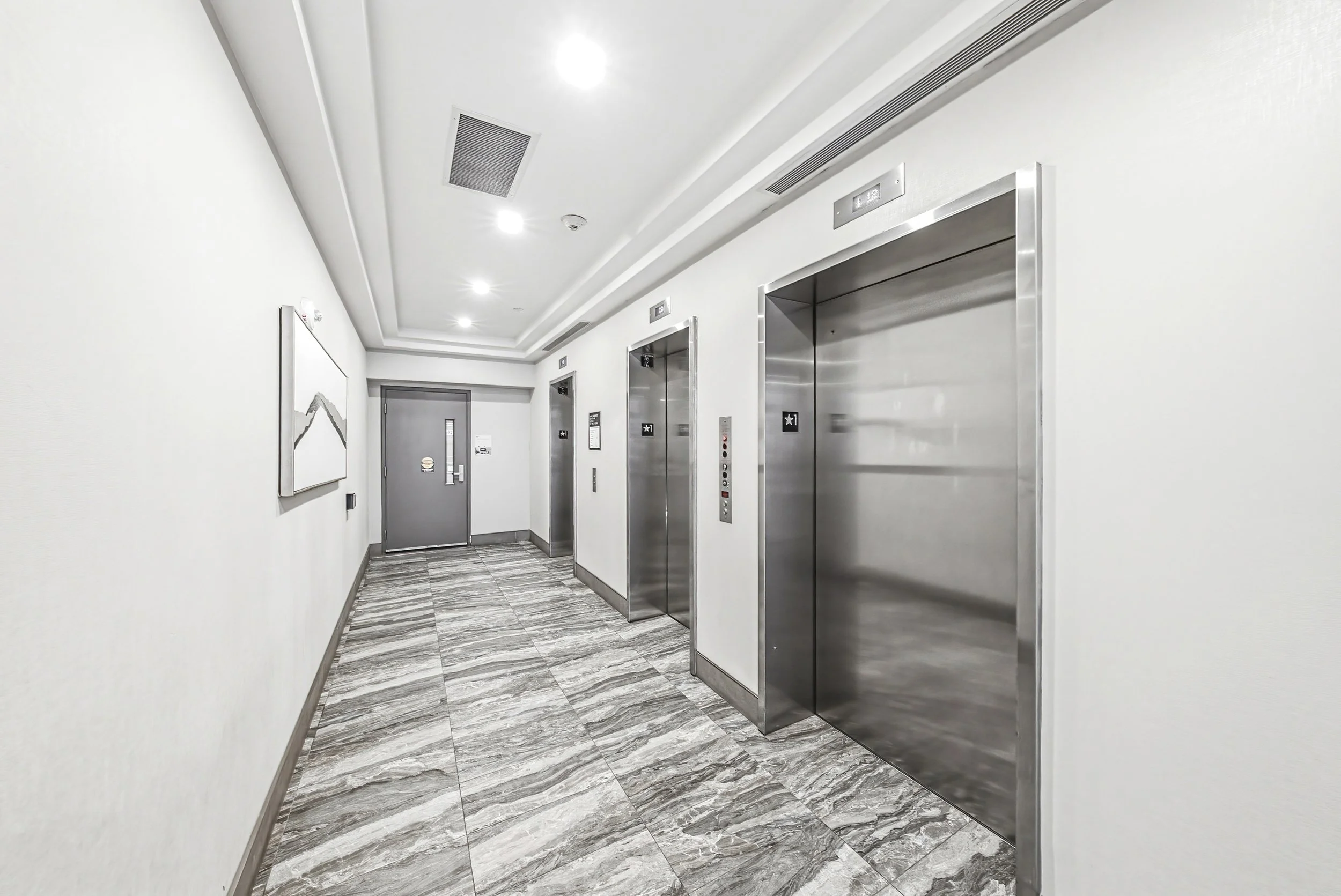 Hallway with two elevator doors, a gray door, and a painting on the wall. The floor is tiled with gray and white marble pattern, and the walls and ceiling are white. There are four ceiling lights and ceiling vents.