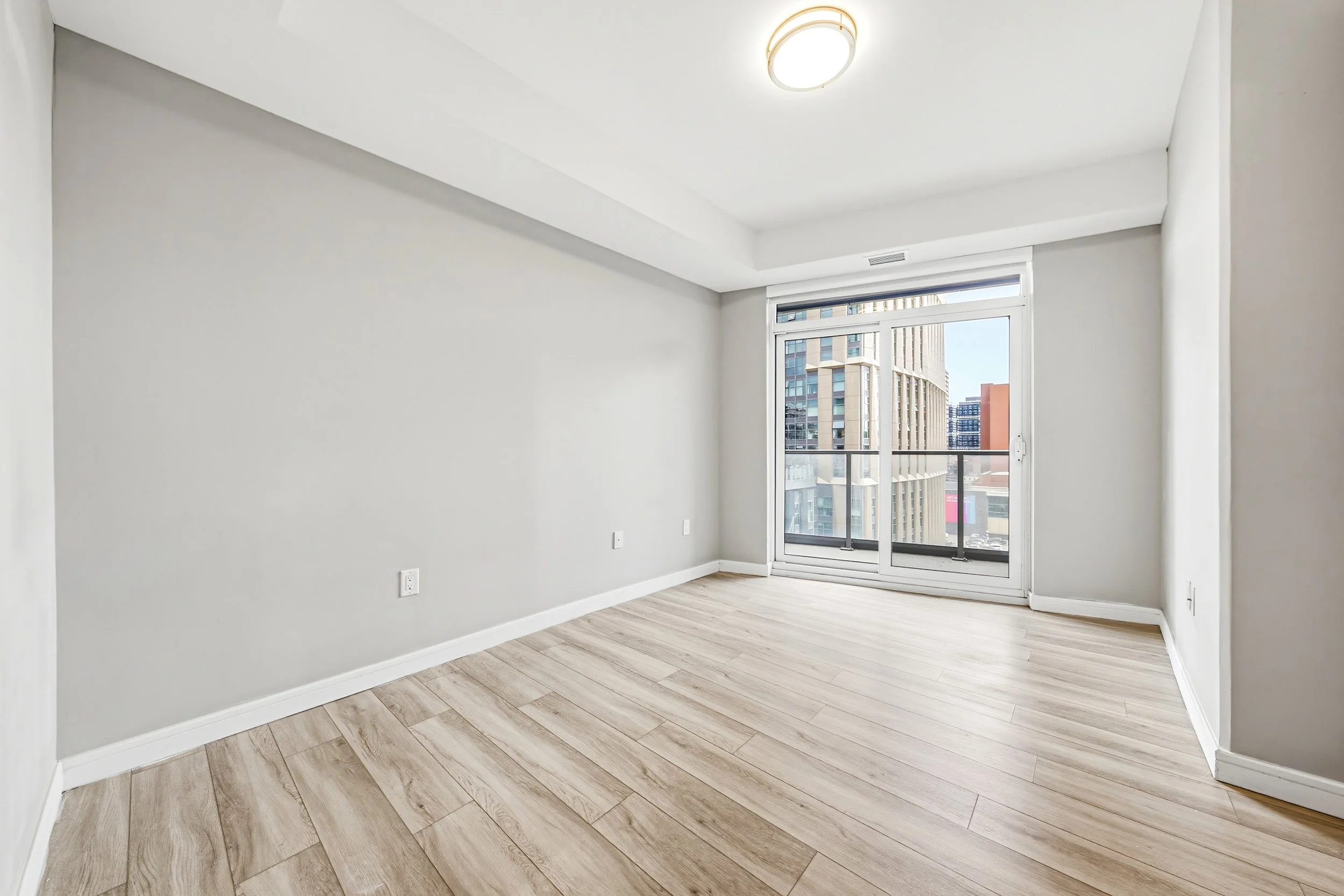 Empty room with light wood flooring, white walls, a large window with a city view, and a ceiling light.
