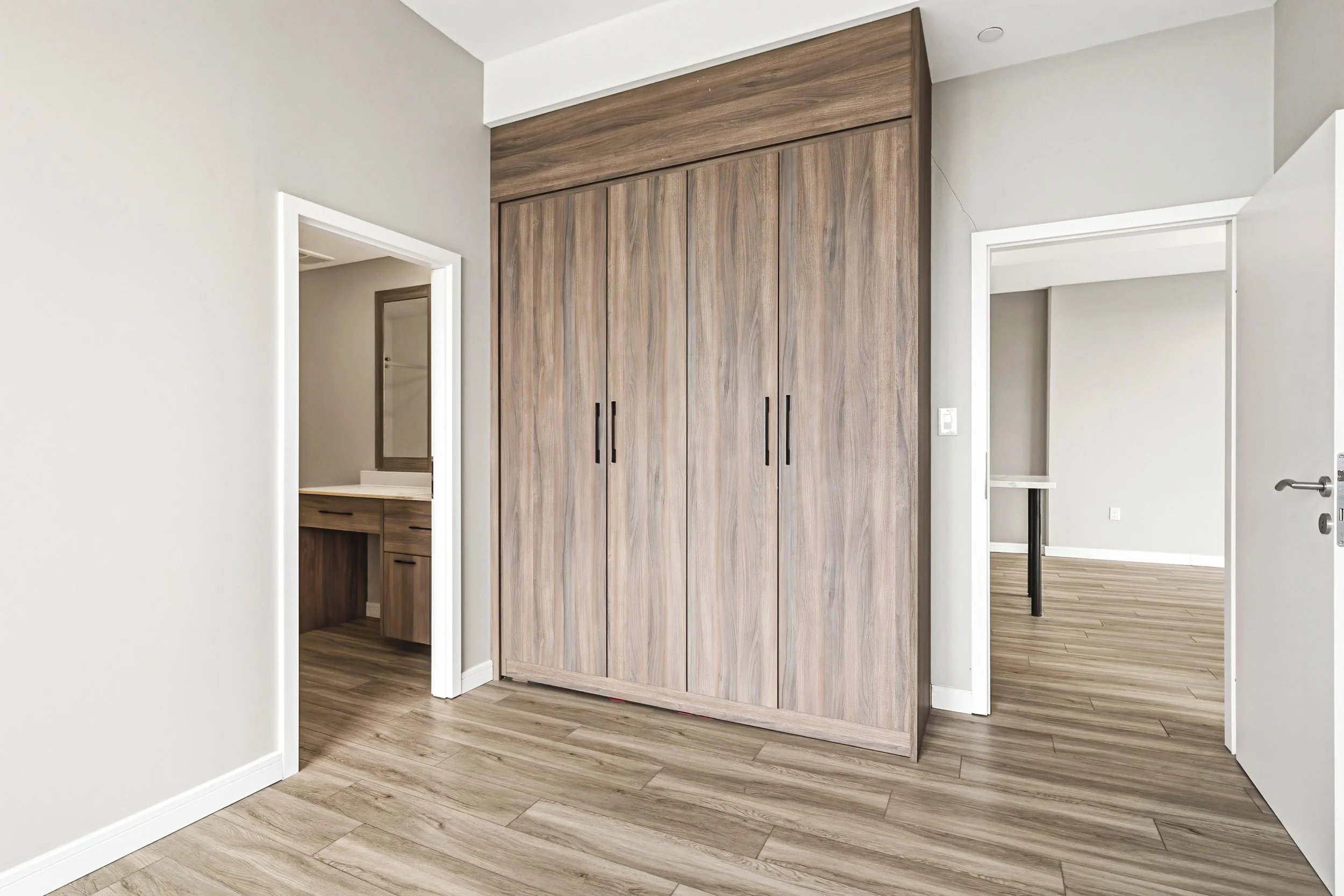 Interior of a modern house with wooden flooring, sliding closet doors, and white walls. Multiple doorways lead to different rooms, including a bathroom with a vanity mirror.
