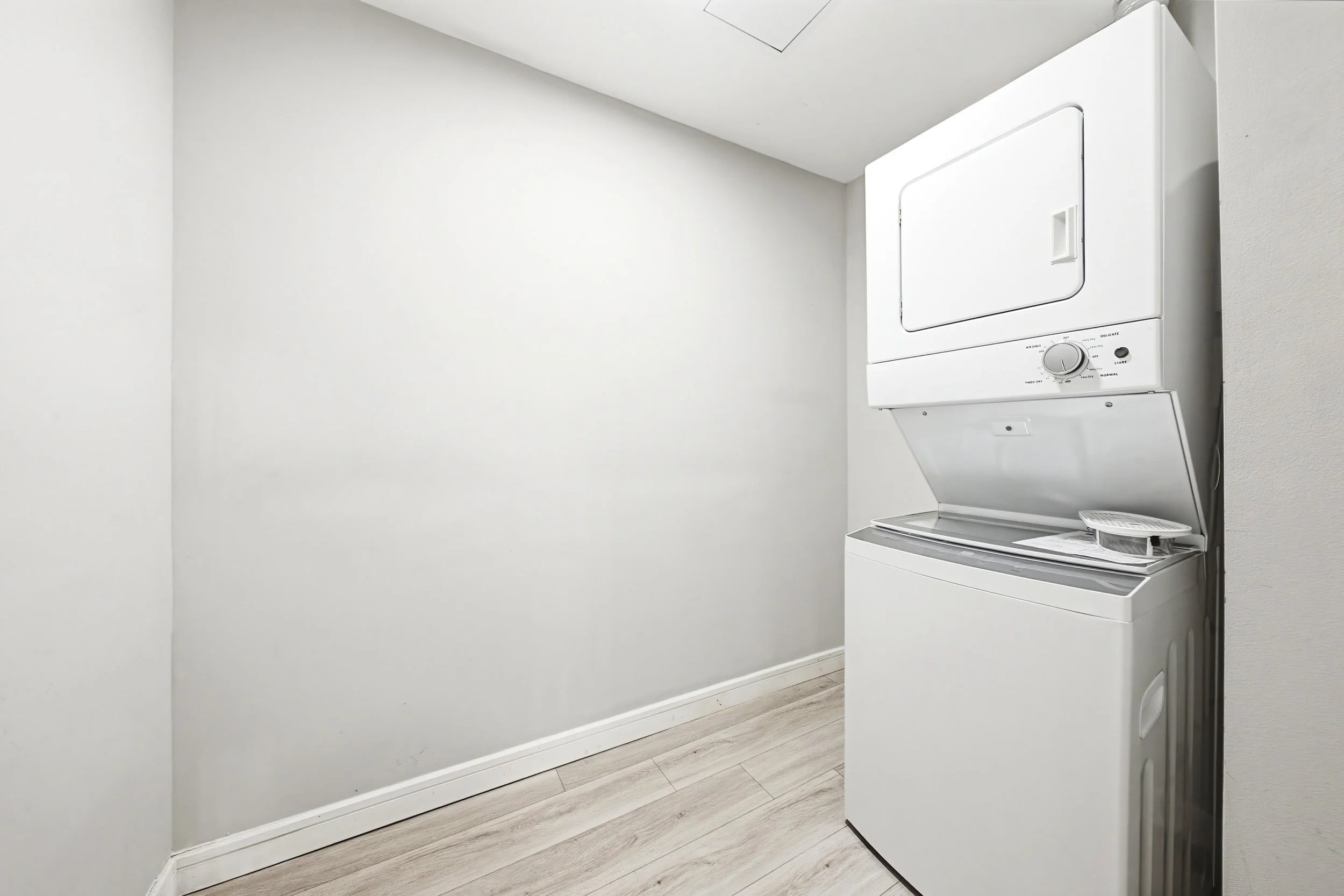 A white stacked washer and dryer unit in a bare laundry room with light-colored wood flooring and plain white walls.