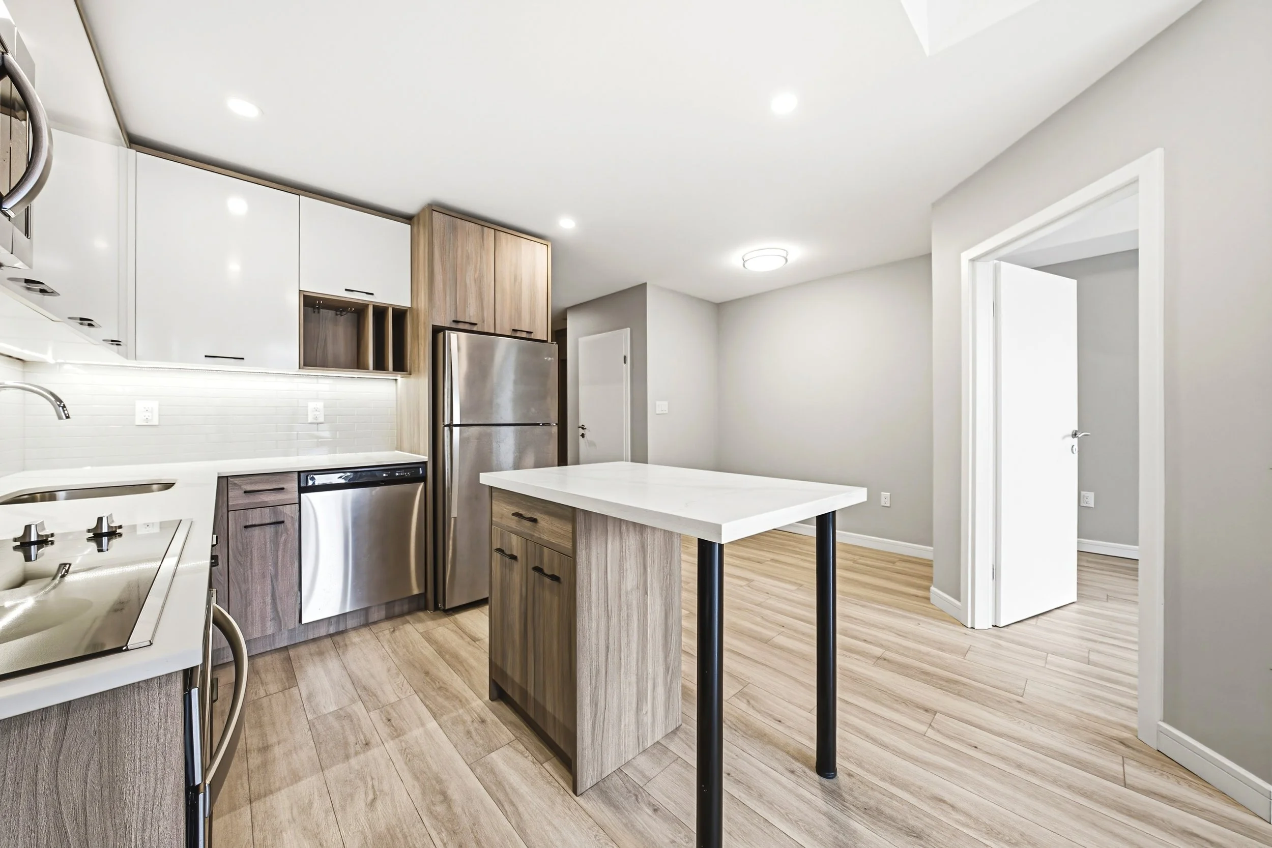 Modern kitchen with white and wood cabinets, stainless steel refrigerator, small kitchen island, light wood flooring, and neutral walls.