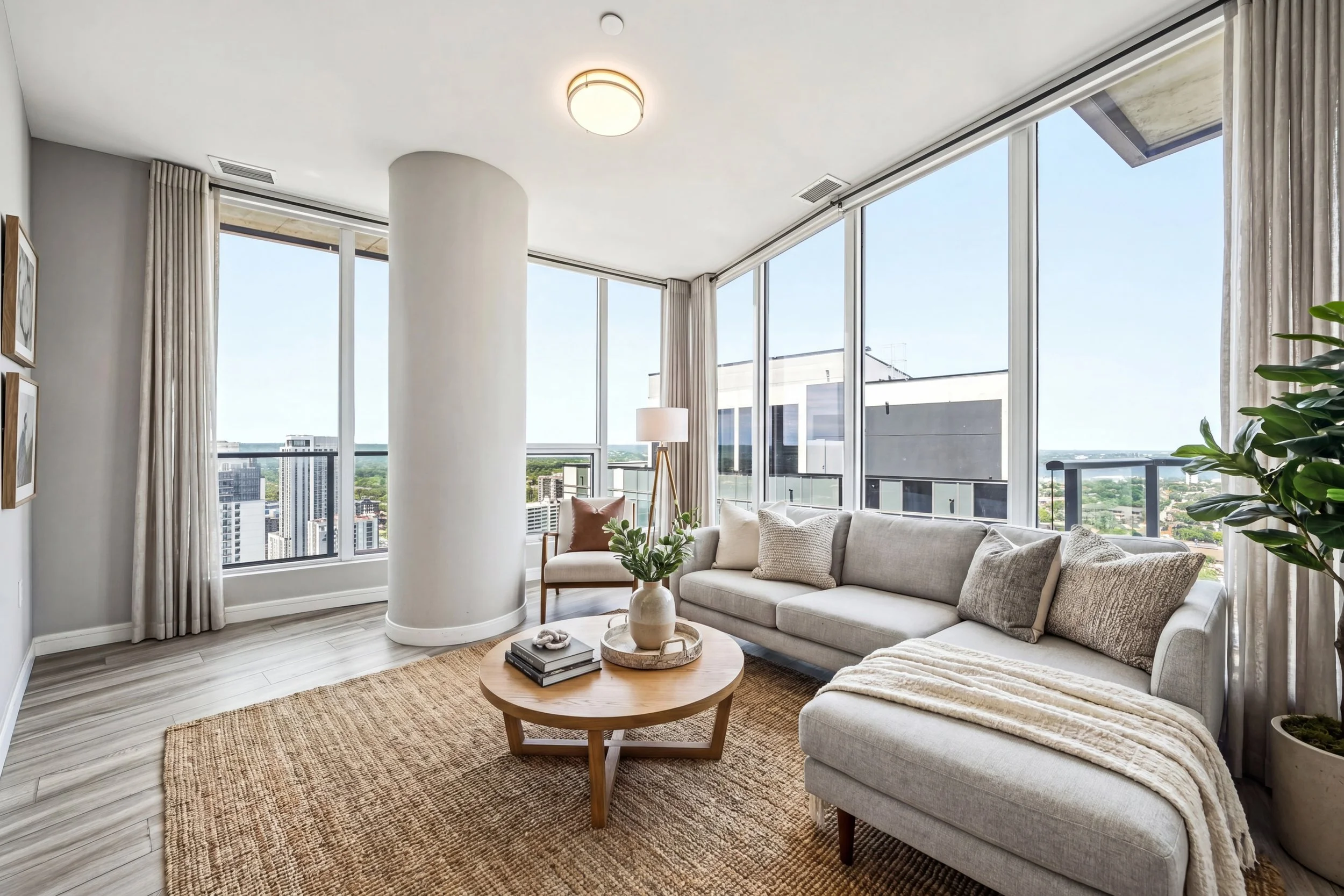 Living room with large windows, beige sofa with pillows, wooden coffee table with books and decorations, light-colored curtains, potted plant, view of cityscape outside.