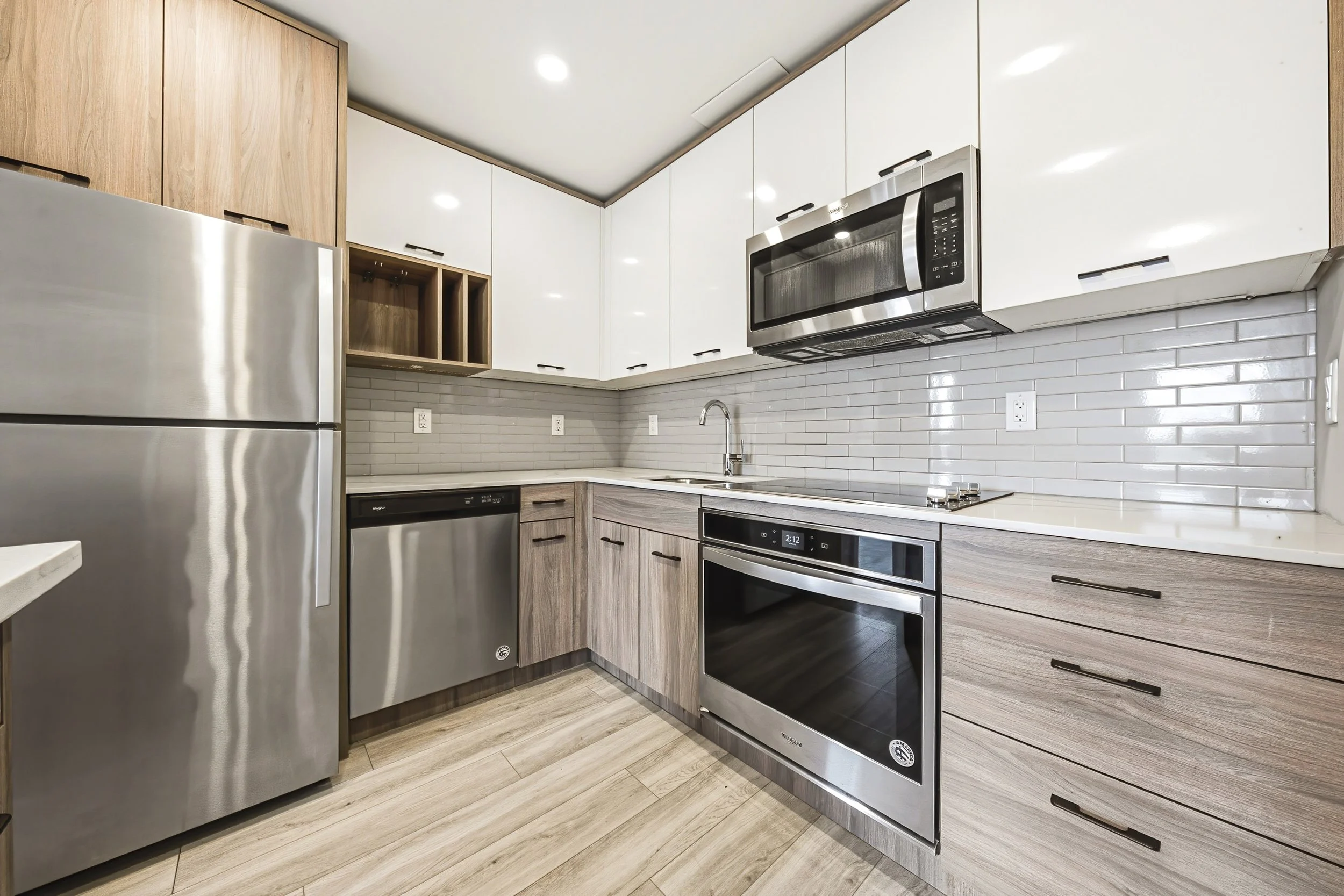 Modern kitchen with white upper cabinets, wood lower cabinets, stainless steel appliances including a microwave, oven, and refrigerator, tile backsplash, and wood-look flooring.