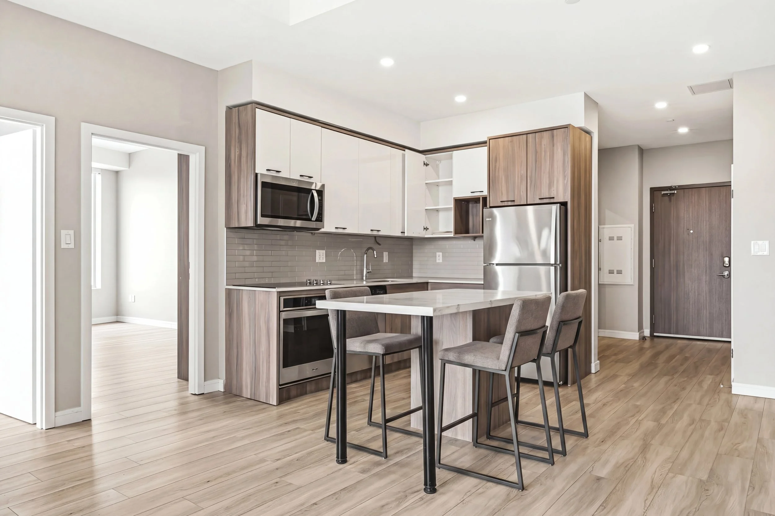 Modern kitchen and dining area with white cabinets, stainless steel microwave and fridge, grey brick backsplash, and light wood flooring.