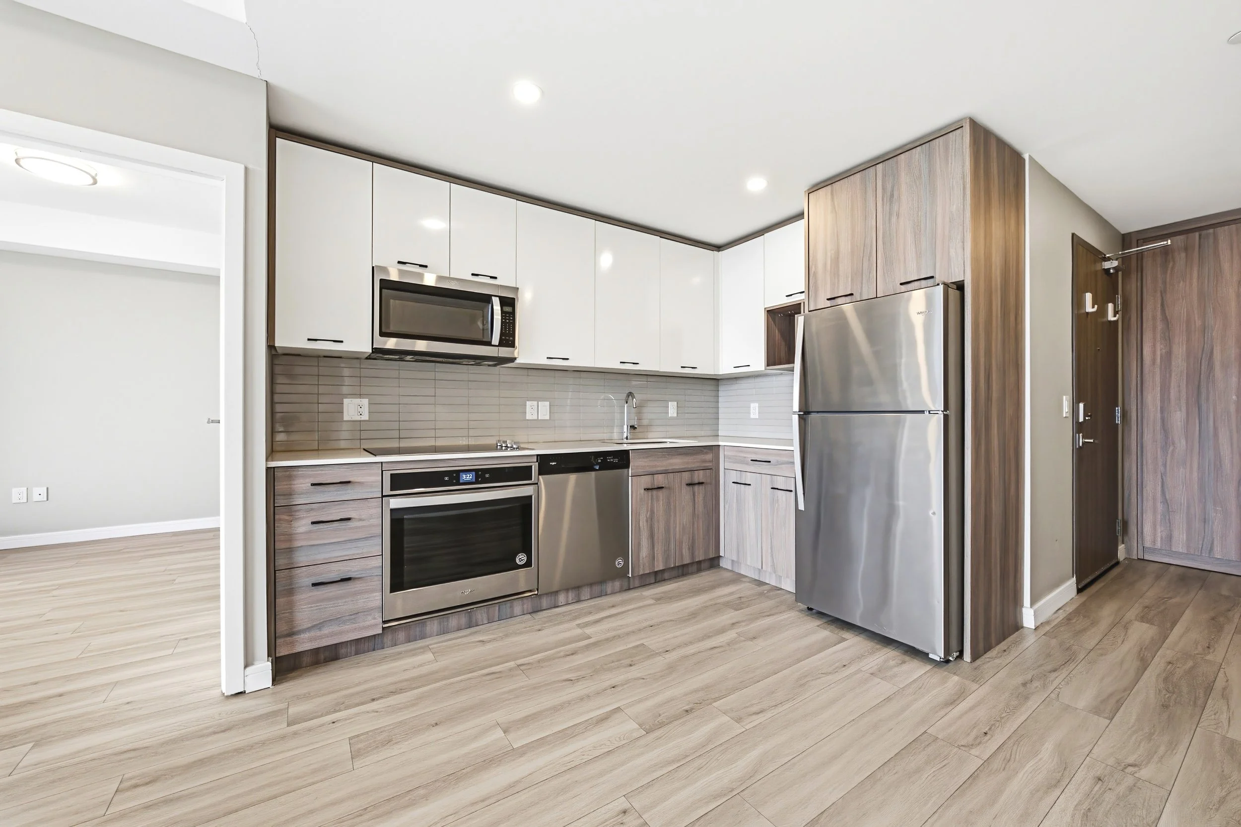 Modern kitchen with white upper cabinets, wooden lower cabinets, stainless steel appliances including a microwave, oven, refrigerator, and dishwasher. Light wood flooring and a white ceiling with recessed lighting.