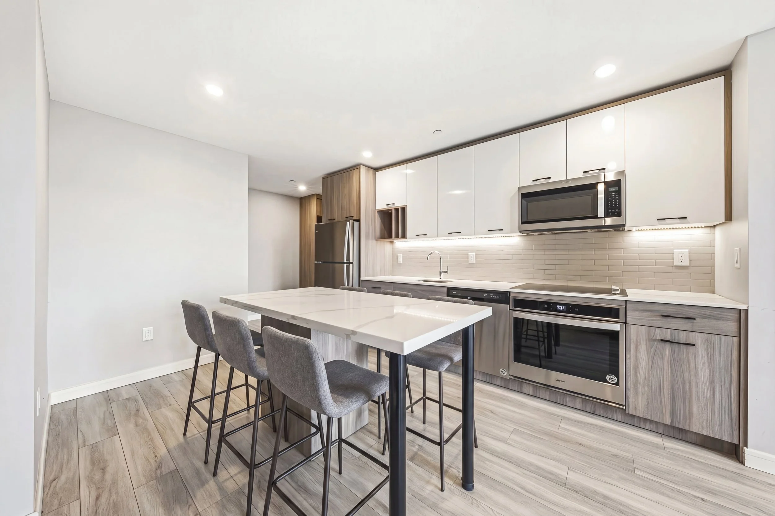 Modern kitchen with white cabinets, stainless steel microwave, oven, and refrigerator, light wood flooring, and a white kitchen island with gray chairs.