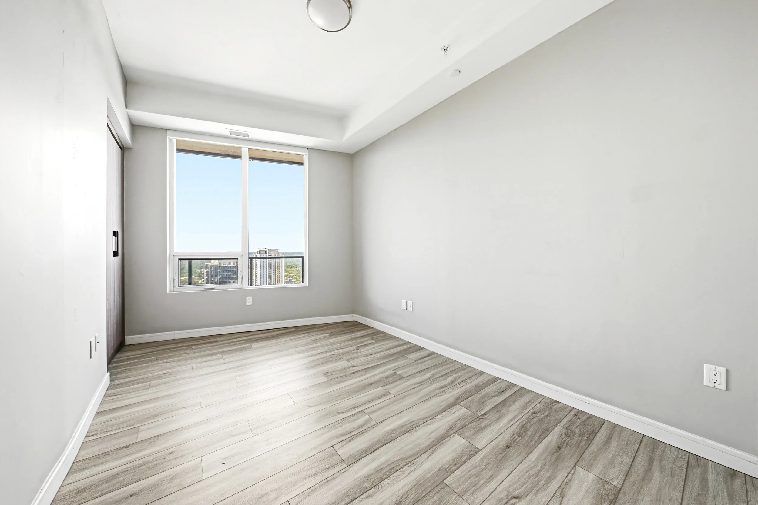 Empty room with large window showing cityscape, white walls, wooden laminate flooring, and electrical outlets.