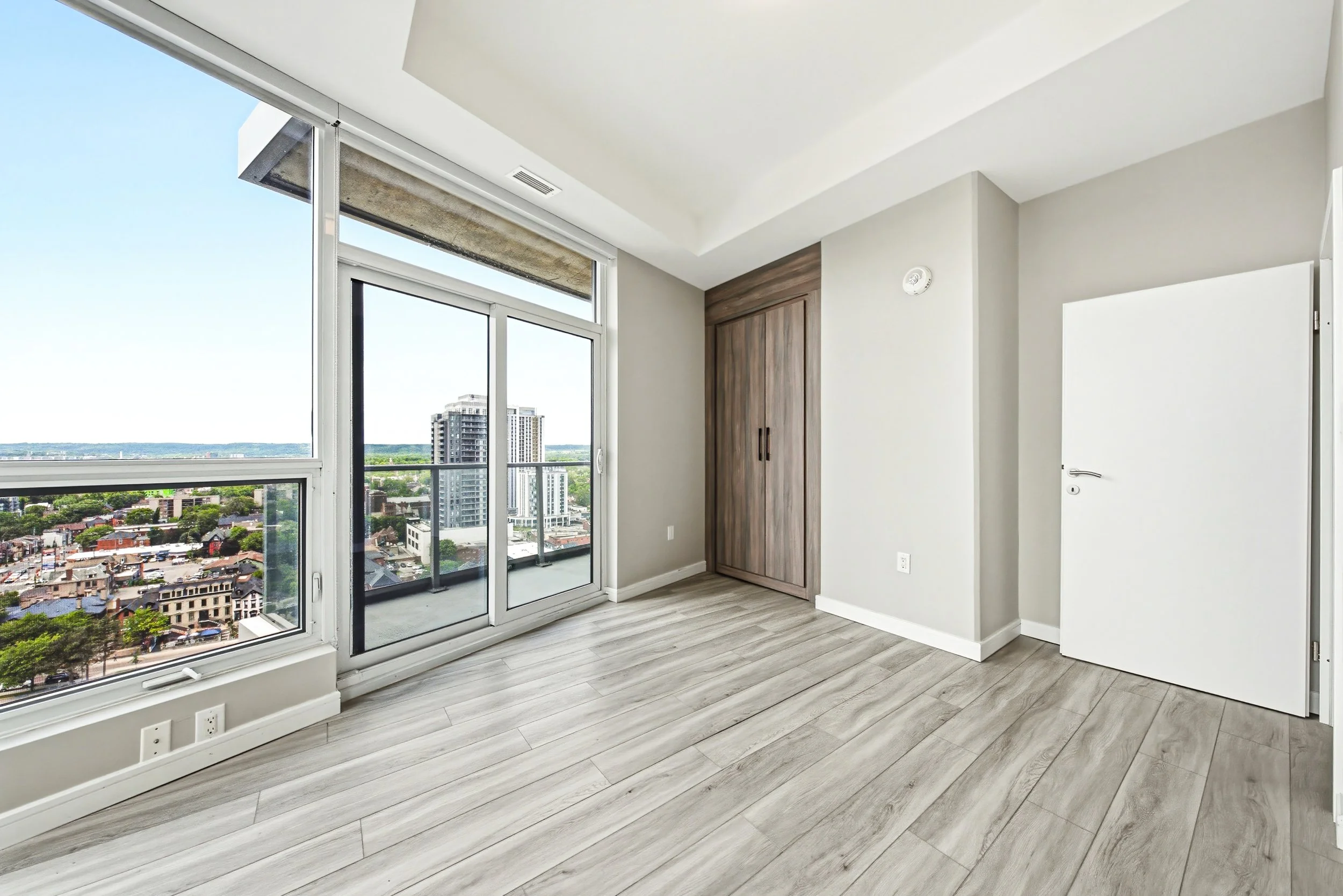 Empty modern room with large windows overlooking a cityscape, white walls, wooden floor, and a closed wooden closet door.
