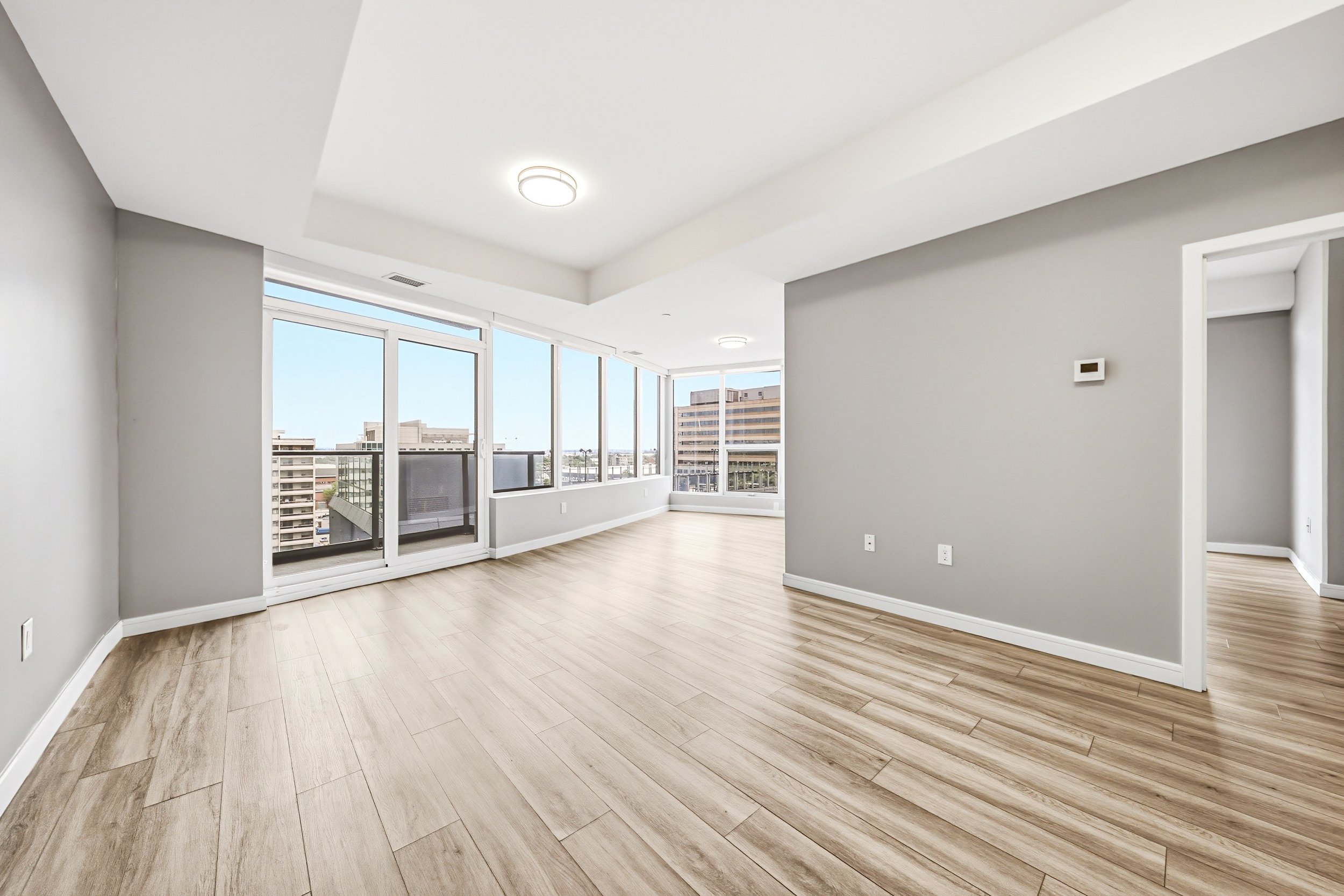Empty living room with large windows, light wood flooring, white walls, and modern ceiling lights.