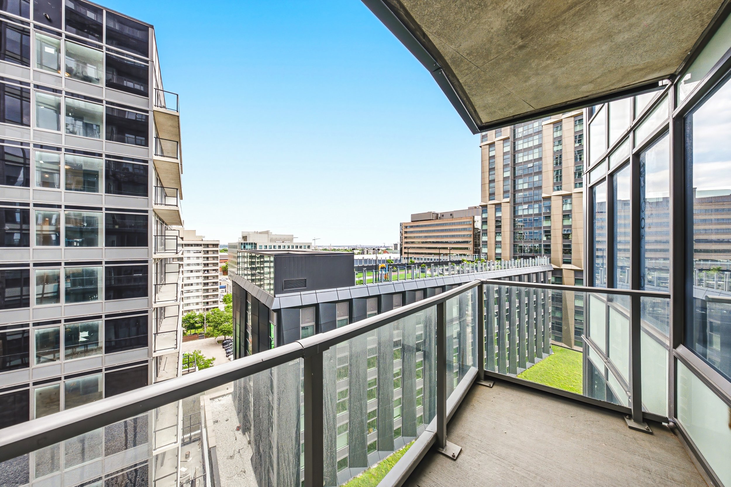 View from a balcony showing neighboring modern apartment buildings with glass exteriors and a green rooftop area, under a clear blue sky.