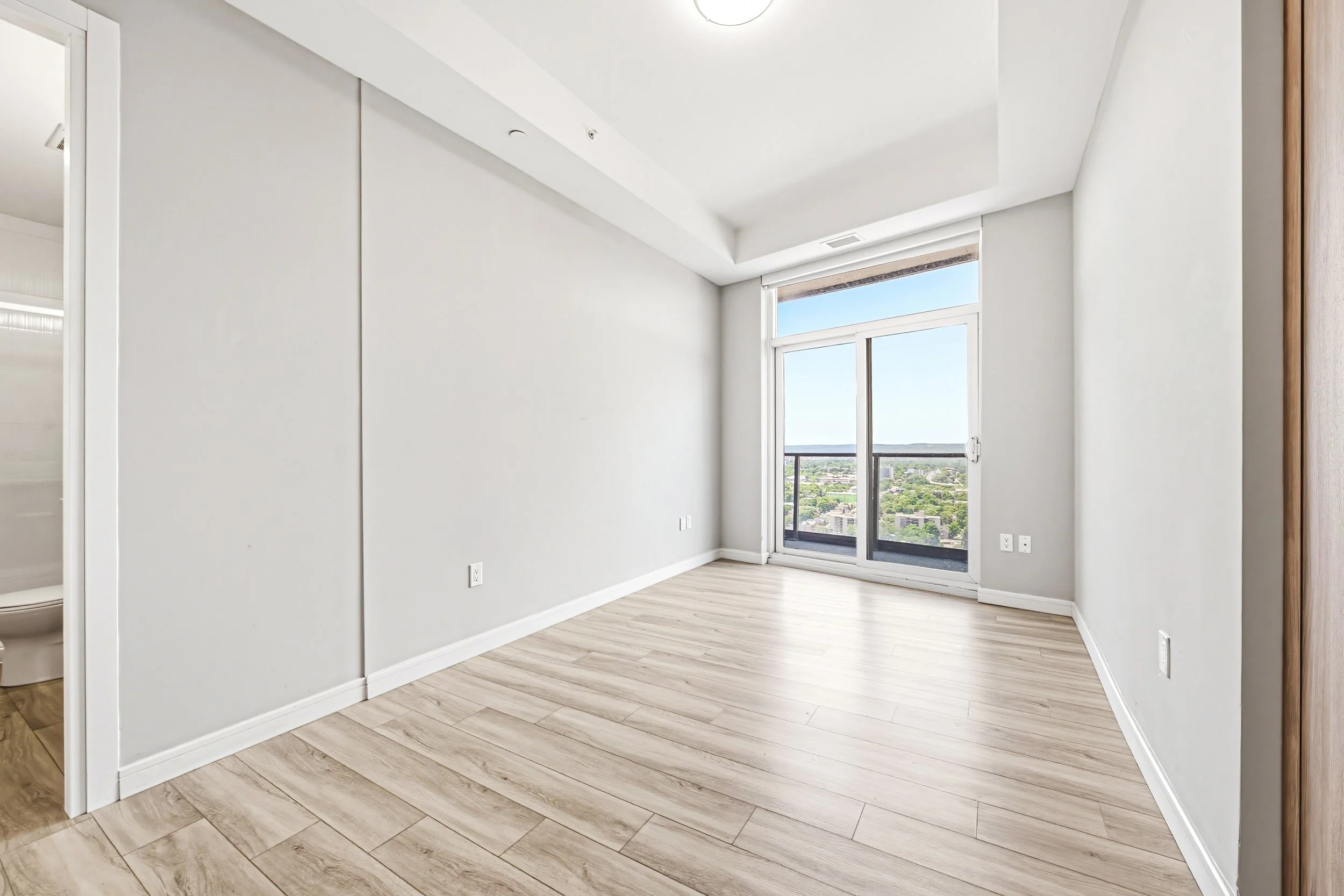 Empty room with light wood floors, white walls, large window with city view, and a sliding glass door leading to a balcony.