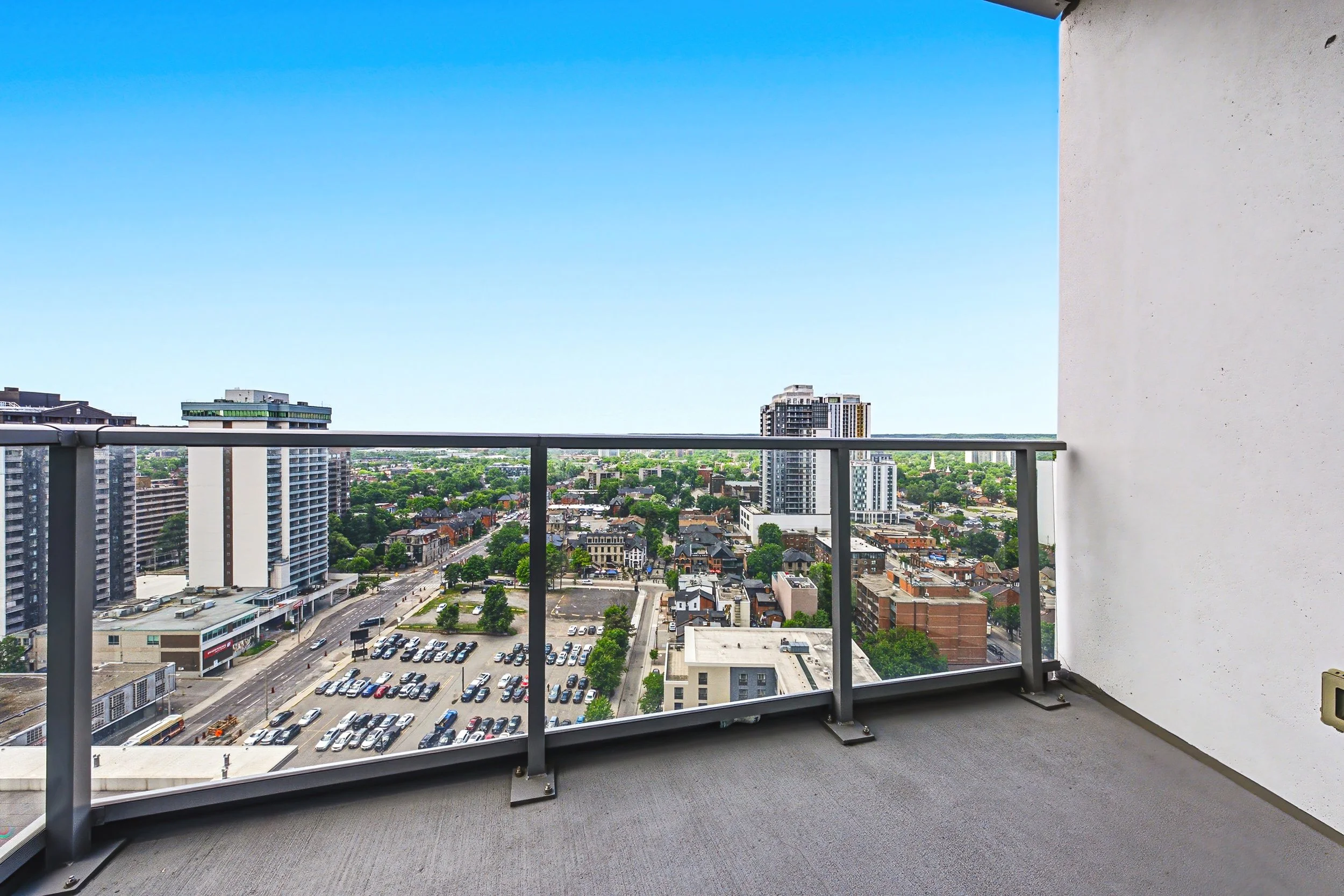 View from a high-rise balcony showing a city skyline with tall buildings, a parking lot, and a street with cars, under a bright blue sky.