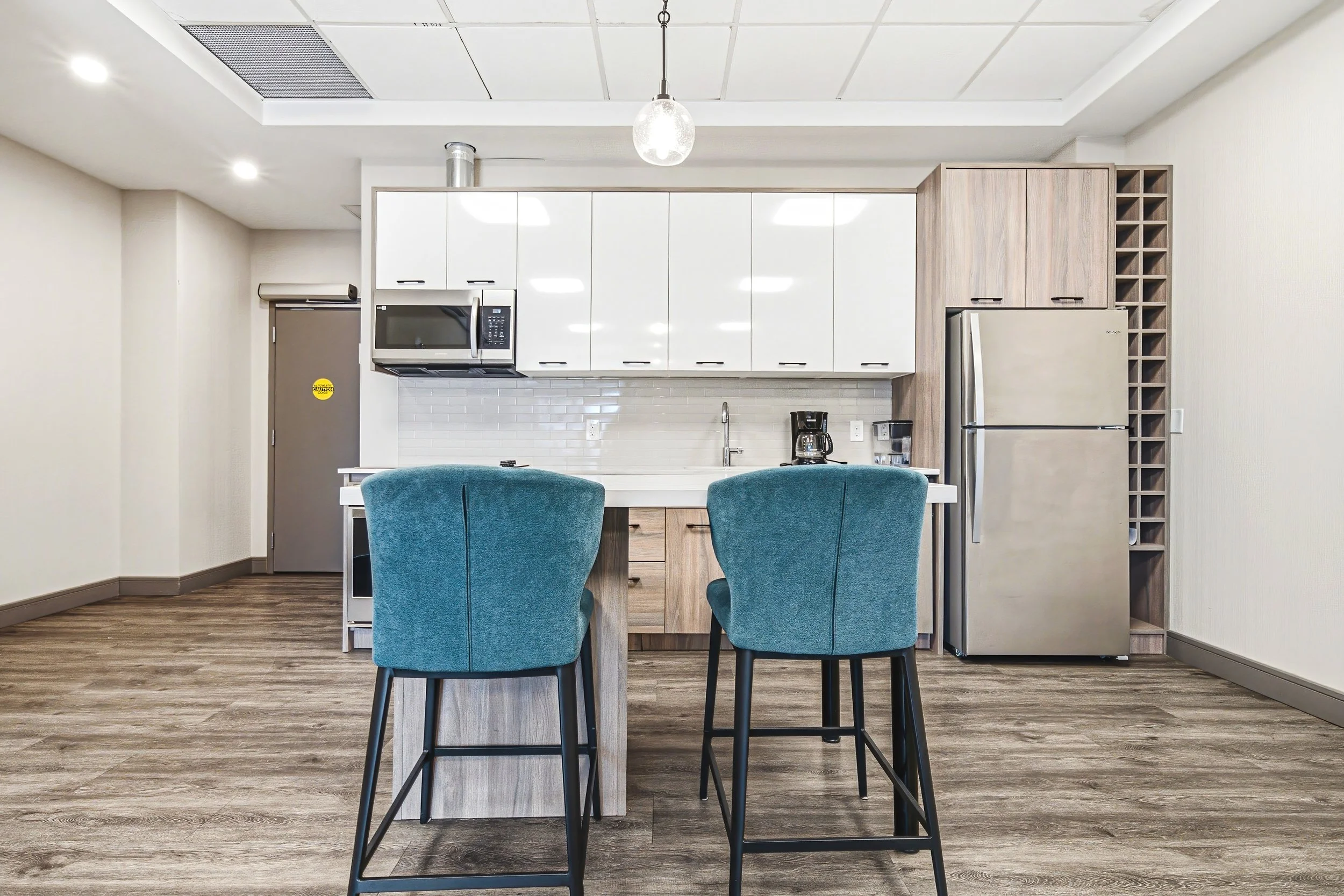 A modern kitchen with white cabinets, a microwave, a refrigerator, and a wine rack. There is a kitchen island with two blue upholstered chairs. The kitchen has wood flooring and a ceiling with recessed lighting and a hanging light fixture.
