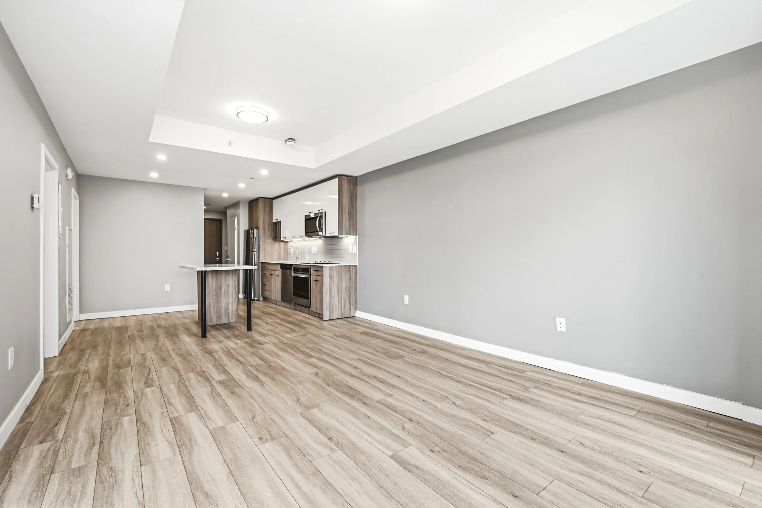 Empty living space with light wood flooring, gray walls, and a modern open kitchen with stainless steel appliances.