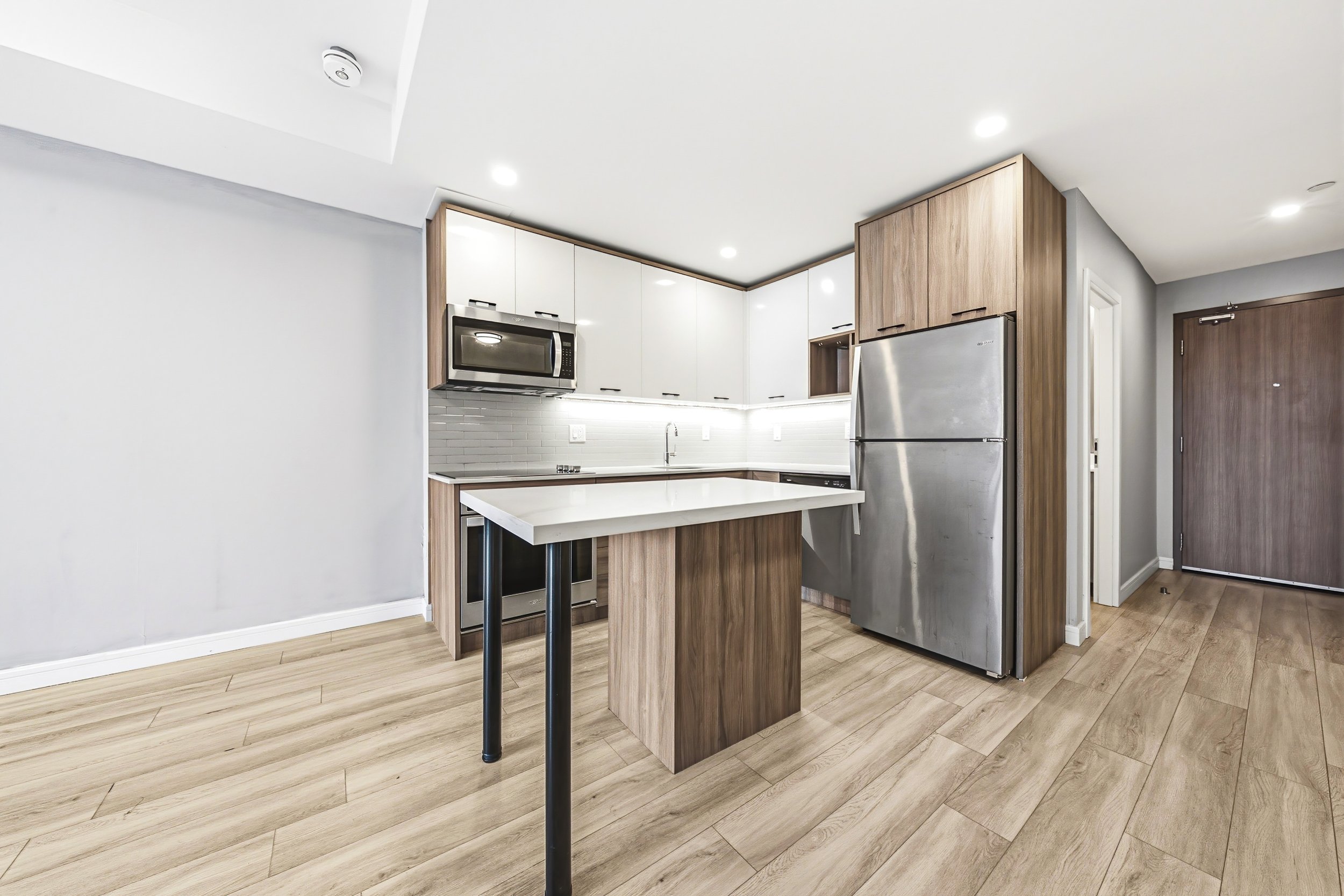 Modern kitchen with white upper cabinets, wooden lower cabinets, stainless steel appliances, light wood flooring, and a small island with a black leg.