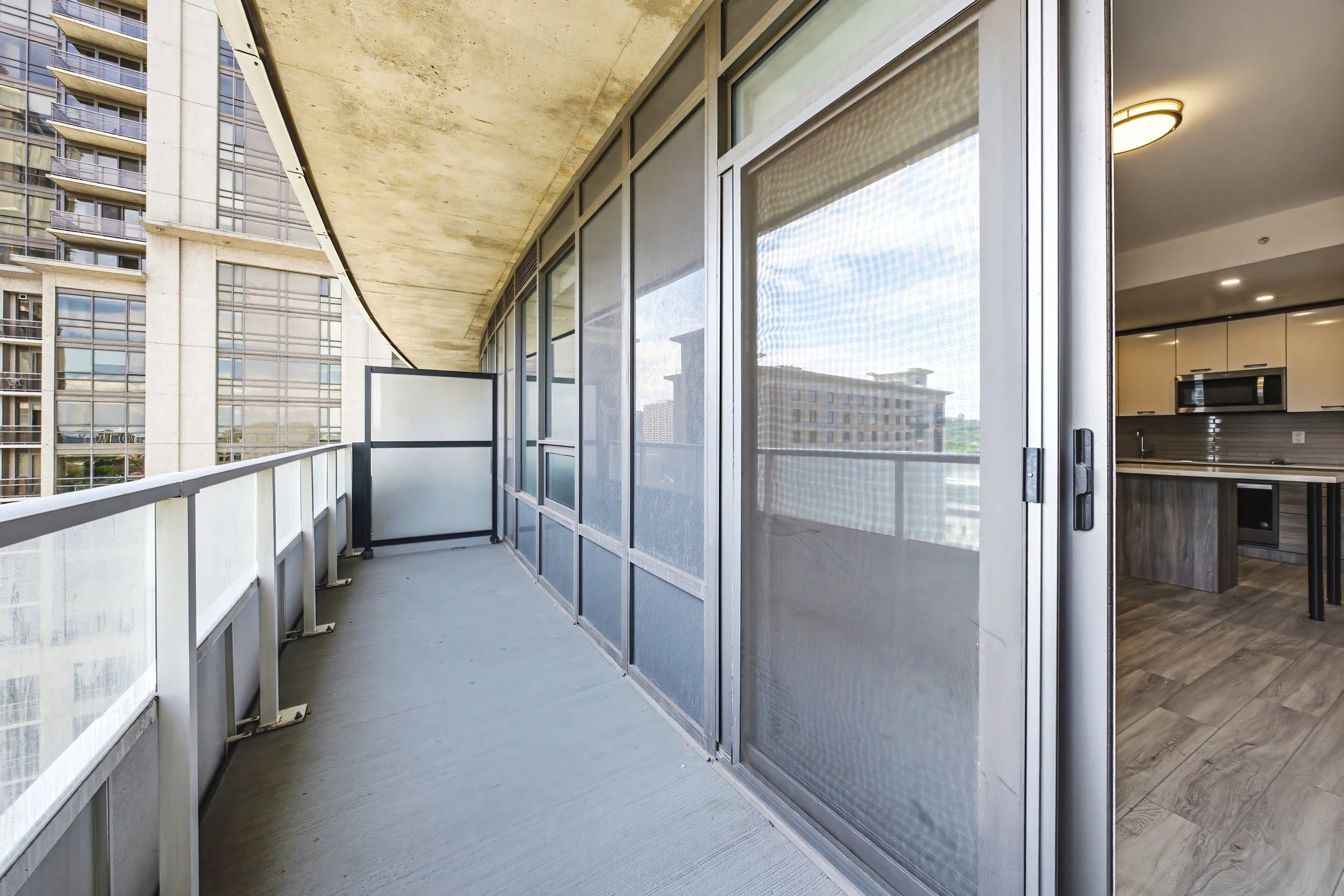 A balcony view with glass sliding doors leading to indoor kitchen area, overlooking neighboring high-rise buildings.