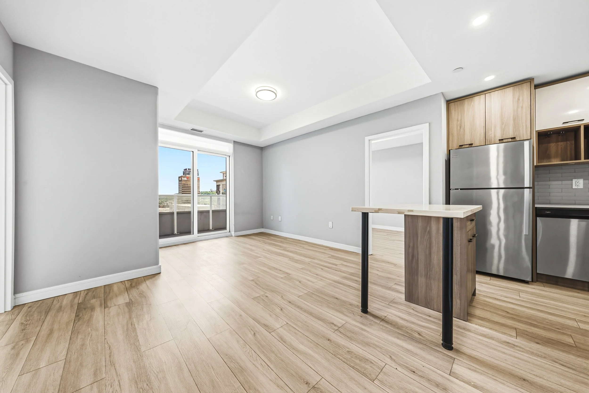 Empty room with light wood flooring, white walls, large windows, a kitchen with wooden cabinets, a stainless steel refrigerator, and a small kitchen island.