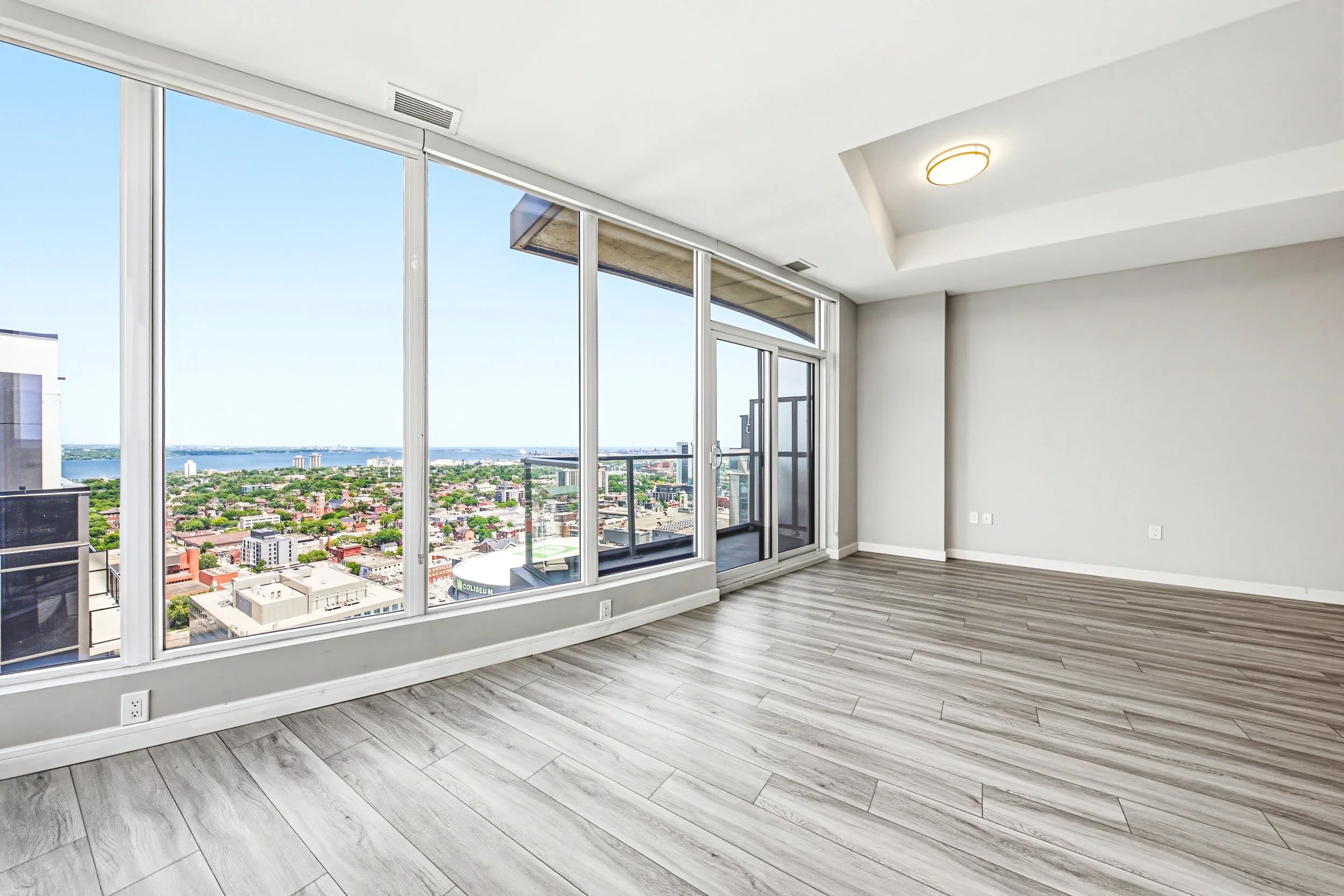 Empty room with large floor-to-ceiling windows showing a city skyline and water view, modern light-colored wood flooring, white walls, and a ceiling with a modern round light fixture.