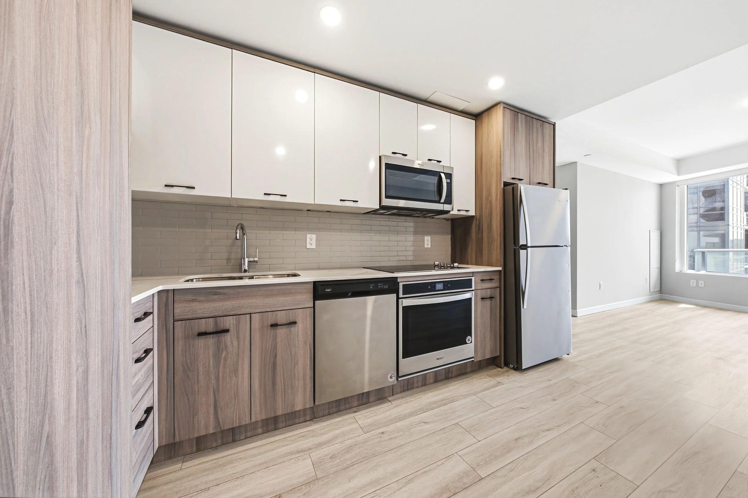 Modern kitchen with white upper cabinets, wood lower cabinets, stainless steel appliances including microwave, oven, and refrigerator, gray brick backsplash, light wood flooring, and large windows letting in natural light.