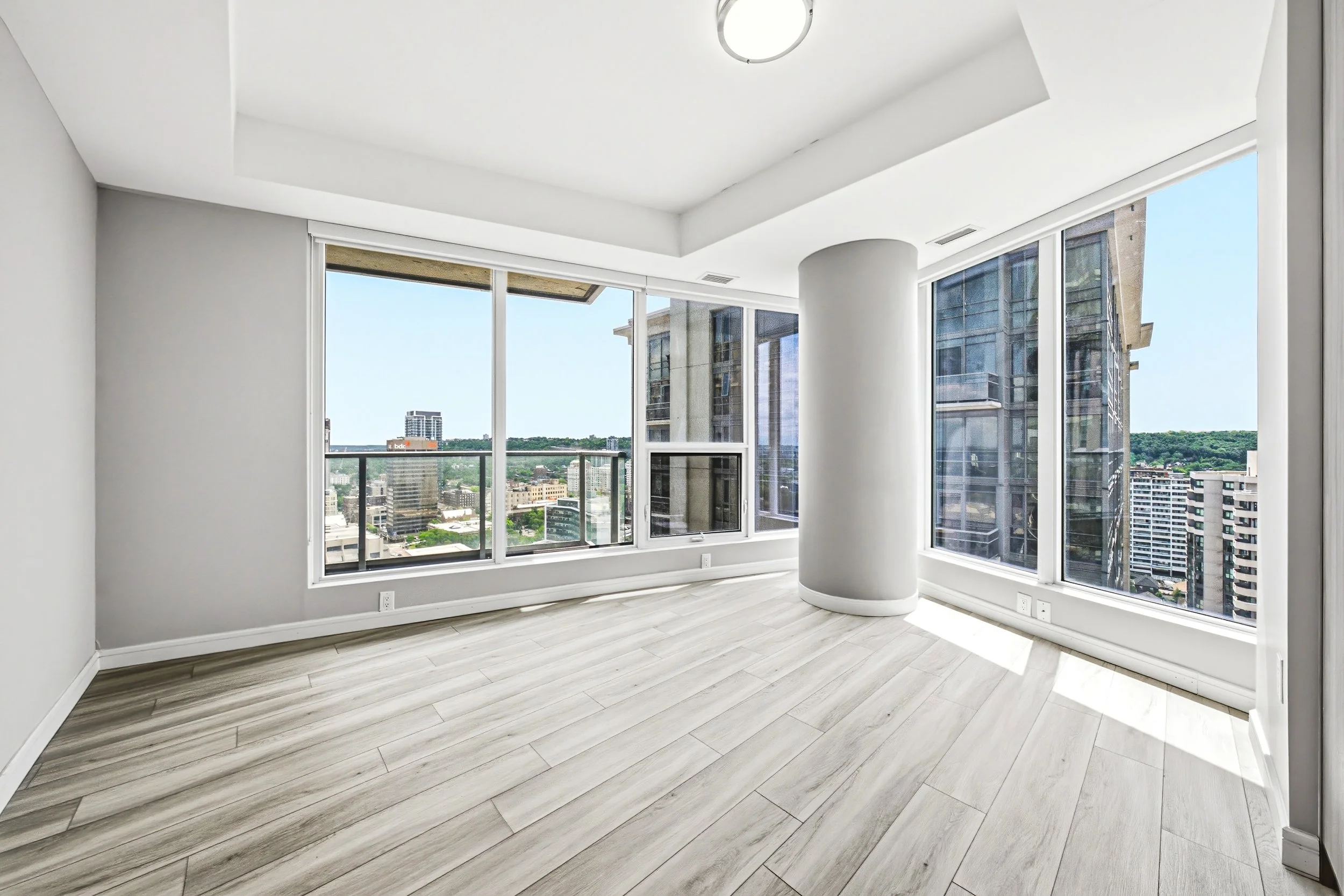 Empty room with large windows and city views, light wood flooring, white walls, and a modern ceiling light fixture.