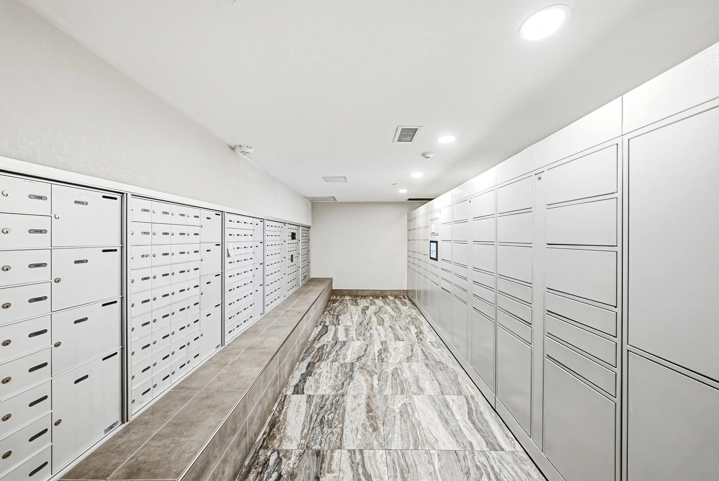 A modern indoor mailroom with white lockers on both sides, a tiered seating area, and marble flooring.