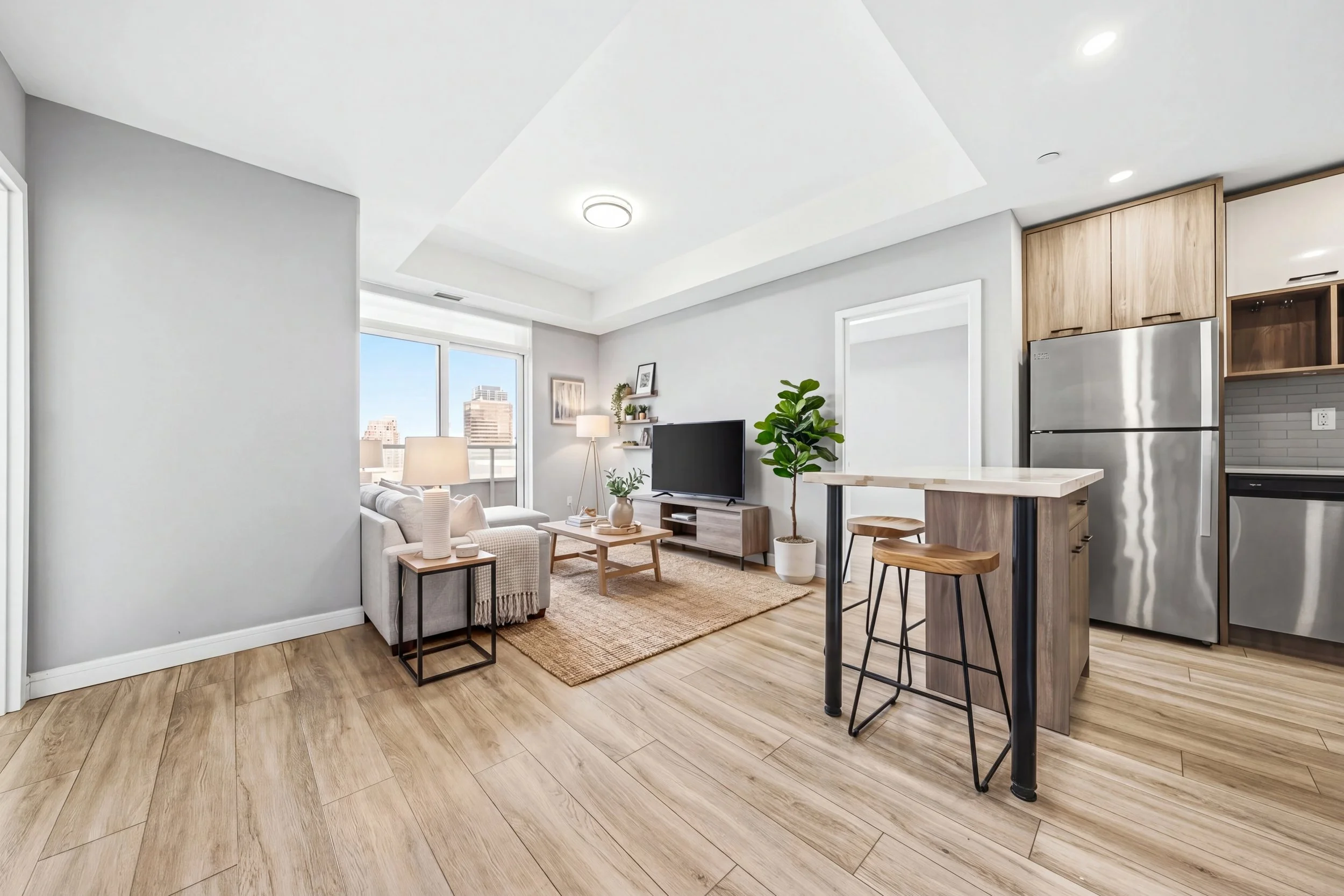 Modern living room with white walls, light wood flooring, large windows with city views, beige sofa, coffee table, TV on a stand, indoor plants, and a kitchen area with a bar counter and wooden cabinets.