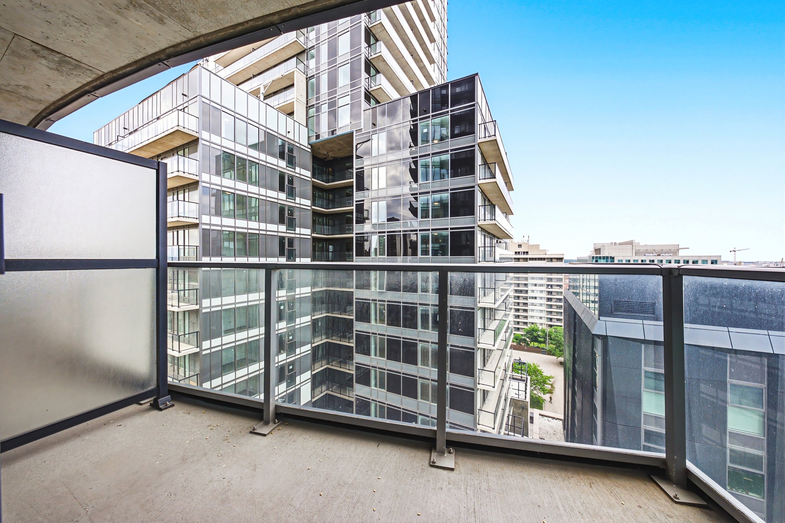 A view from a balcony or terrace of a modern glass high-rise building in an urban area, with other tall buildings and a clear blue sky in the background.