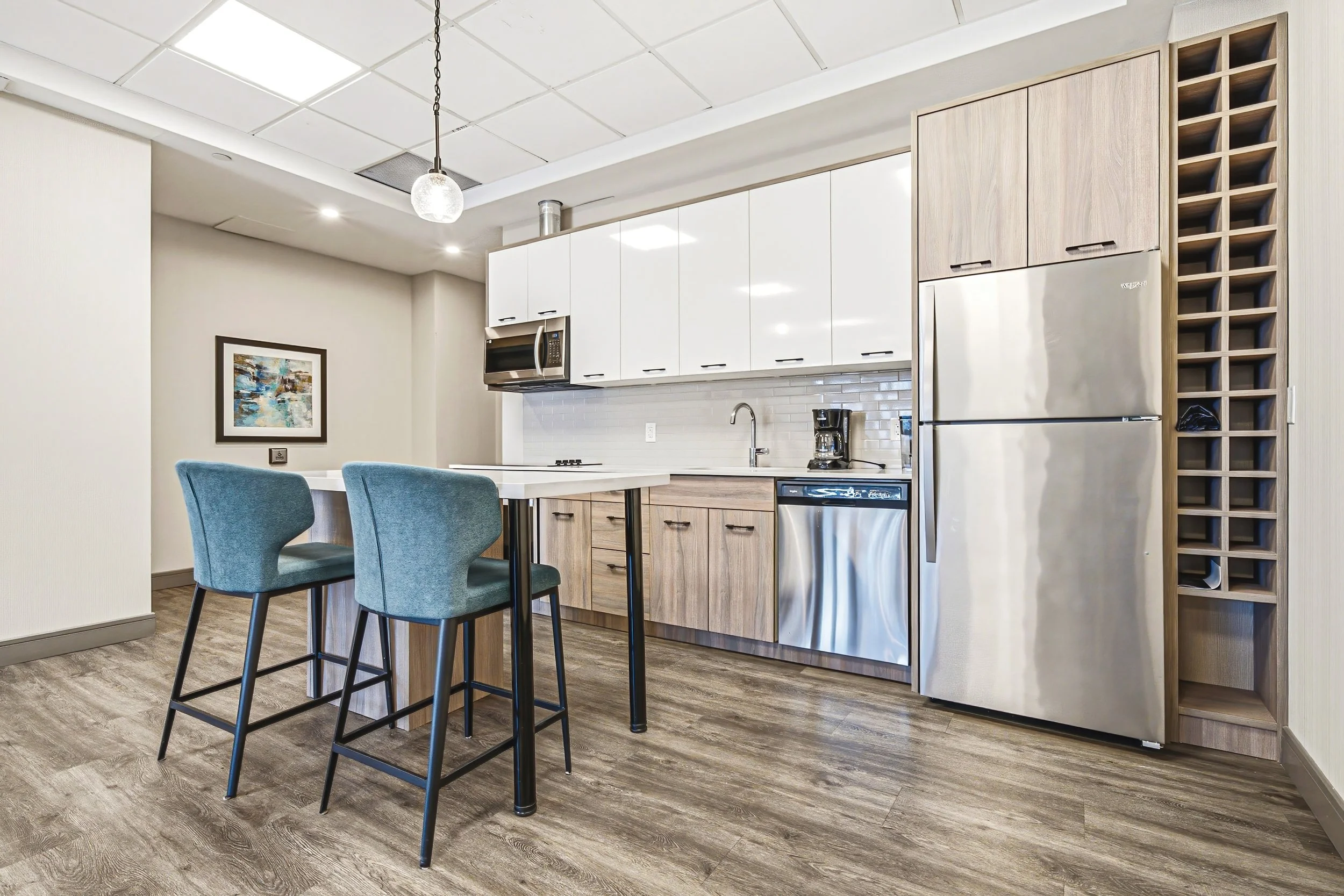 Modern kitchen with white cabinets, wooden drawers, stainless steel refrigerator, and a small dining table with four blue chairs. Artwork on the wall and wood-look flooring.