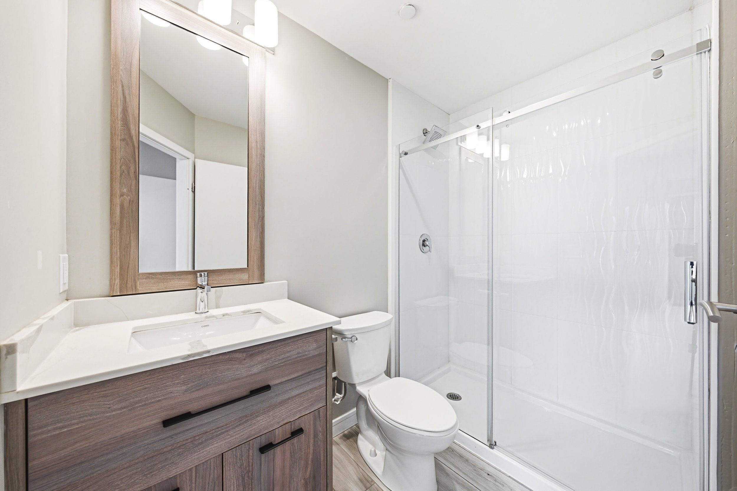 A modern bathroom with a white sink vanity, a large mirror, a toilet, and a glass-enclosed shower with white tiles. The vanity has a wood finish with black drawer pulls, and there is a wooden framed mirror above the sink. The bathroom has neutral-col