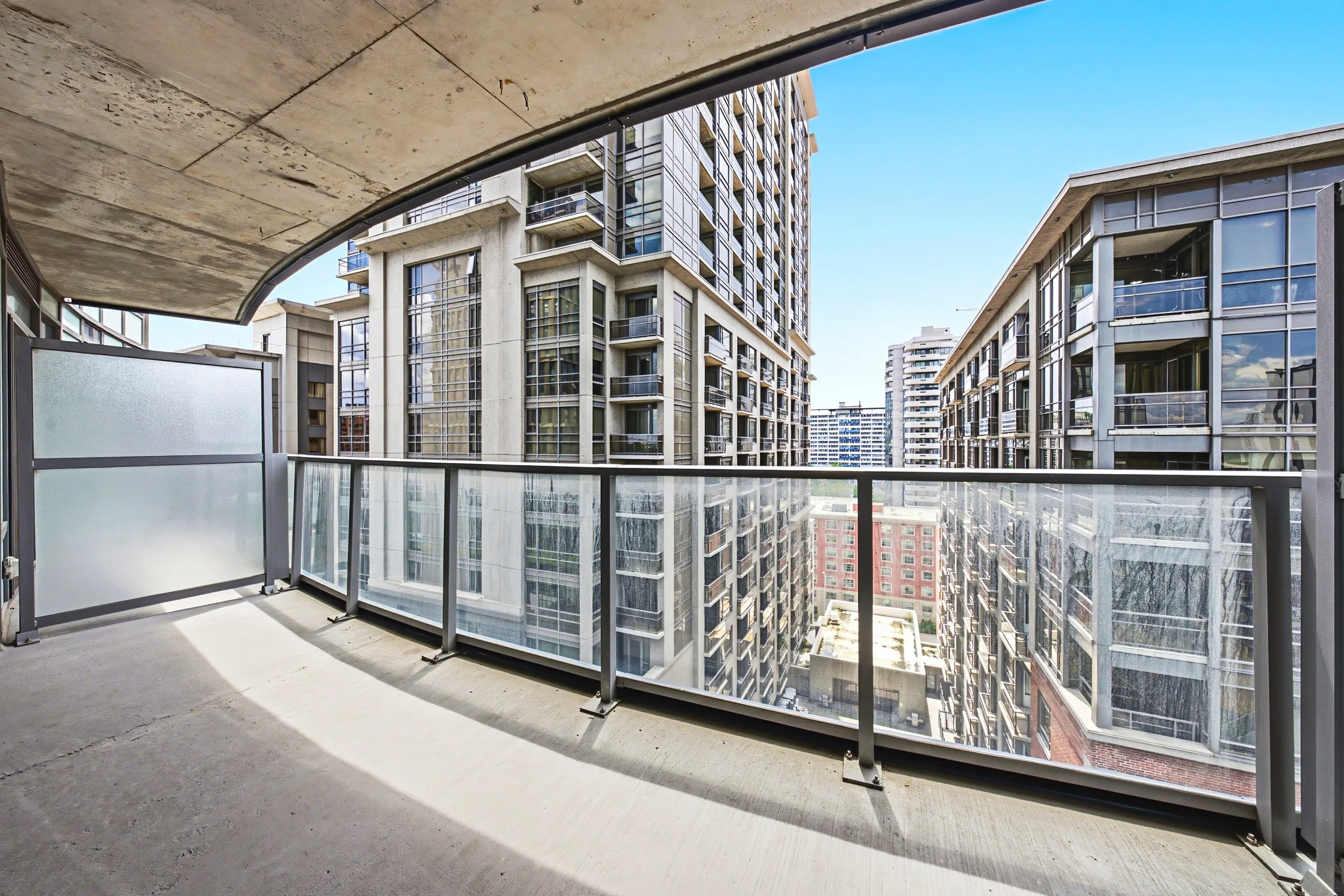 View from a balcony with glass railing overlooking multiple high-rise apartment buildings under a clear blue sky.