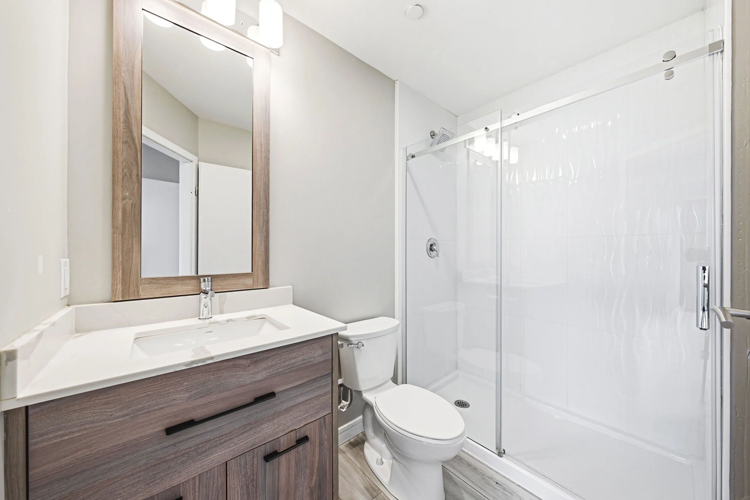 Bathroom with a wooden framed mirror, a white sink with a dark wooden vanity, a white toilet, and a glass shower door. Light-colored walls and wood-look flooring.