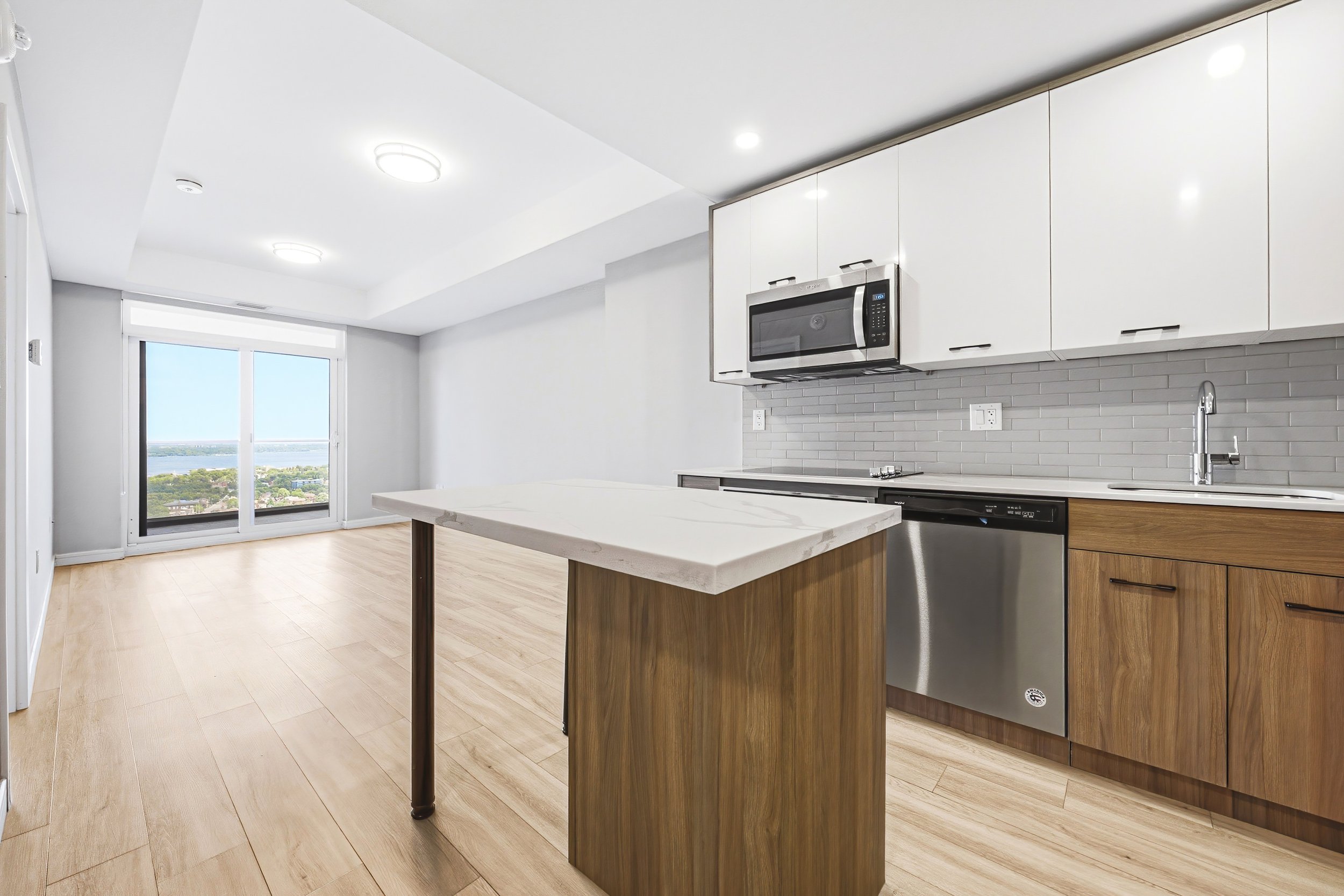 Modern kitchen with white cabinets, a microwave, dishwasher, small kitchen island with a marble top, hardwood floors, large window, and view of trees and water outside.