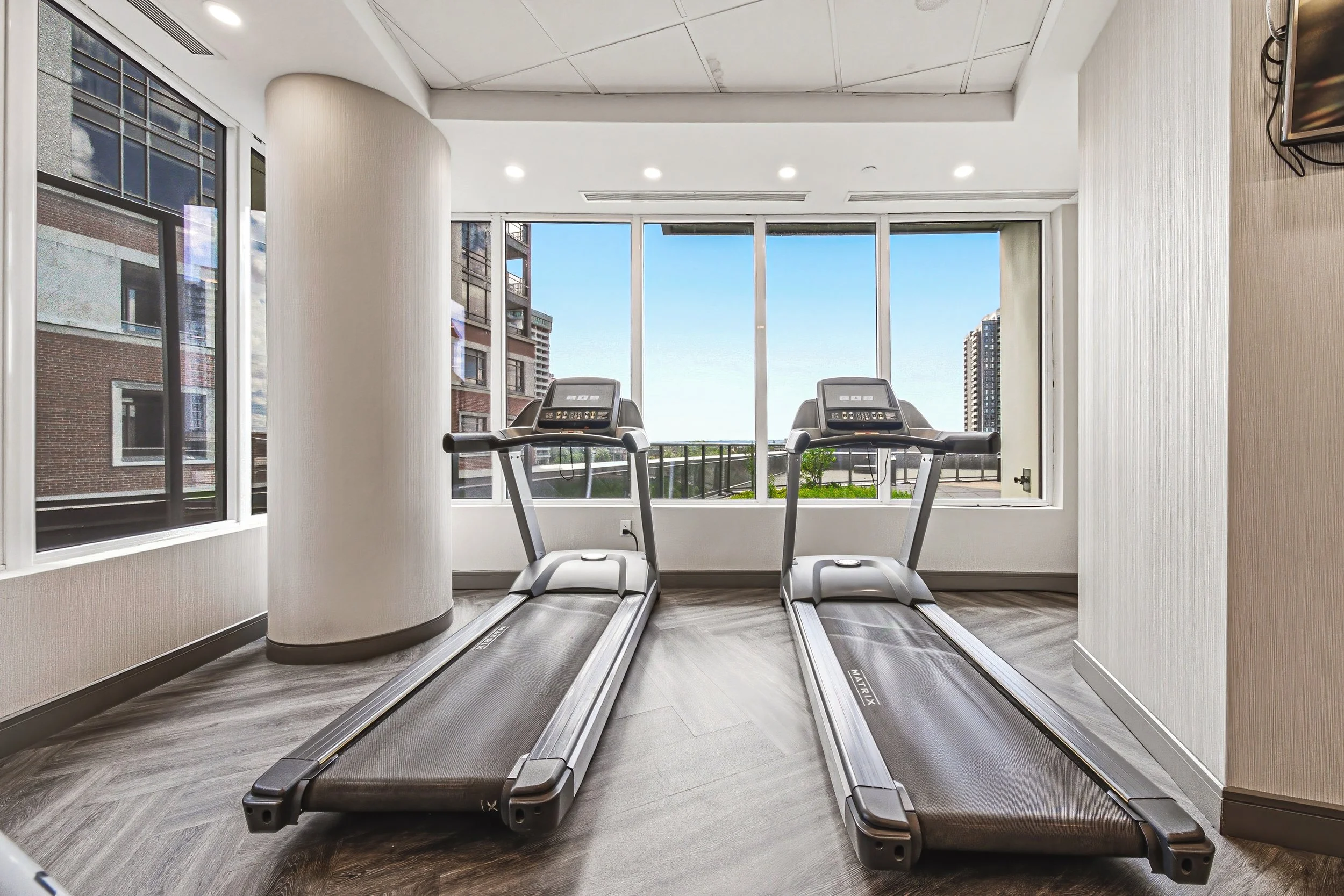 Two treadmill exercise machines facing large windows with city buildings and a blue sky outside in a modern indoor gym.