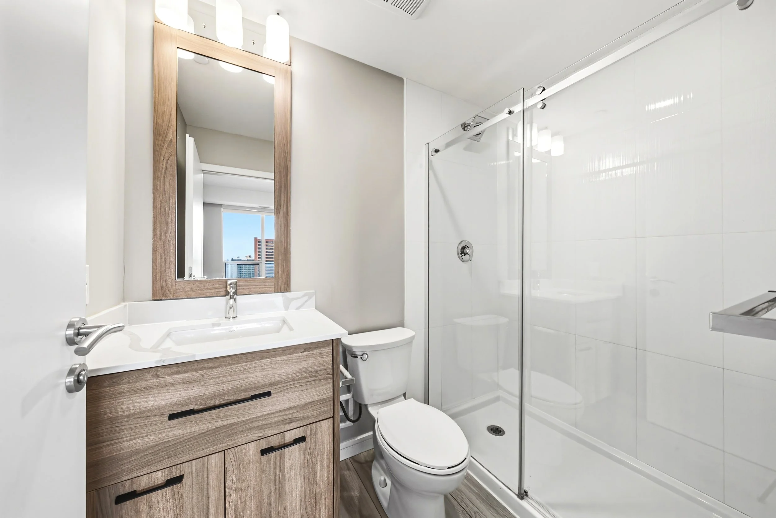 Modern small bathroom with a wood vanity, white sink, and mirror, a toilet, a shower with glass door, and a window showing city buildings outside.