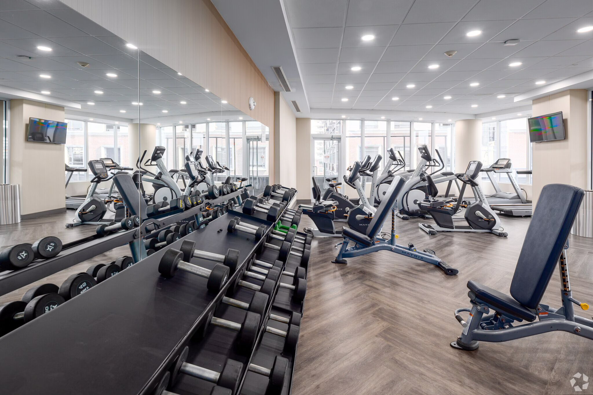 Modern fitness center with treadmills, ellipticals, and free weights, large windows, and ceiling lights.