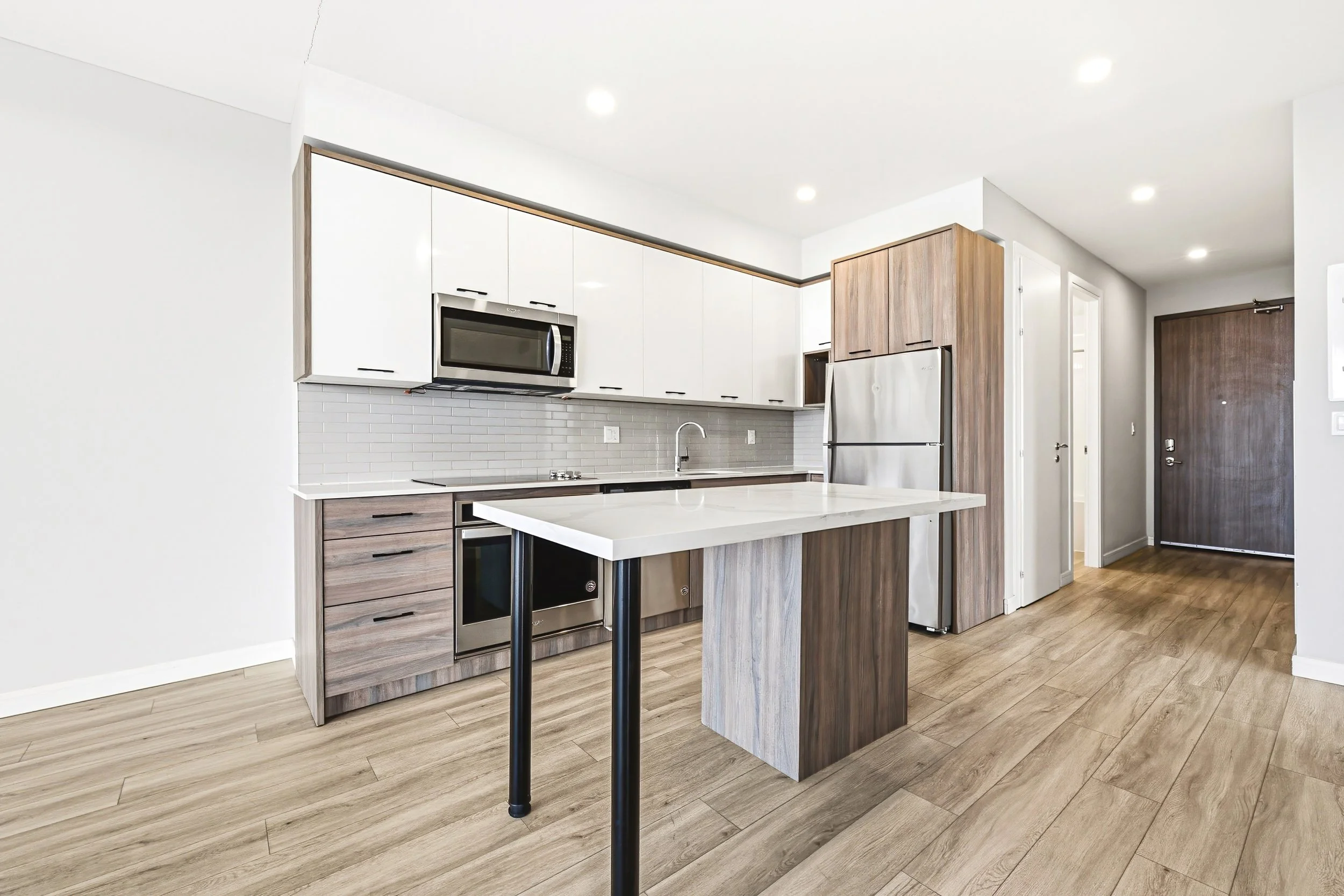 Modern kitchen with white upper cabinets, wood lower cabinets, stainless steel microwave and refrigerator, white countertop island, light wood flooring, and recessed ceiling lights.